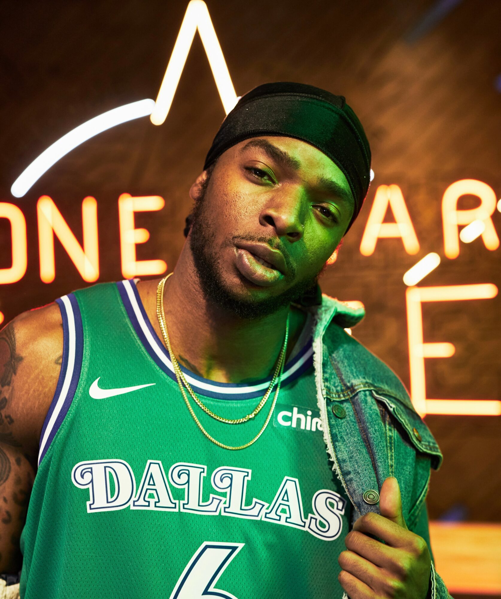Young man wearing a Dallas basketball jersey and a black headband, standing in front of a neon sign, looking at the camera.