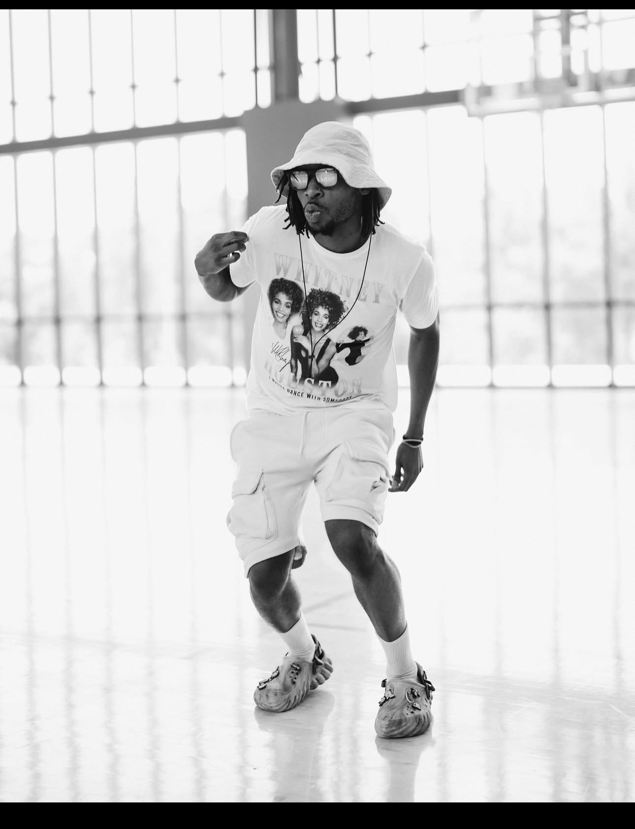 Person with sunglasses, hat, and graphic t-shirt walking indoors, with large windows in the background.