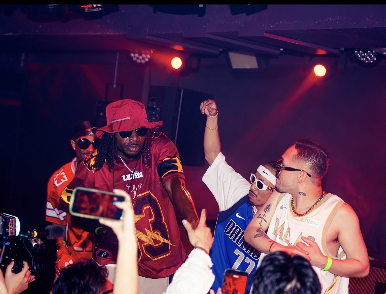 Group of people on stage with colorful lighting, some wearing sunglasses and casual clothing, engaging with the audience.