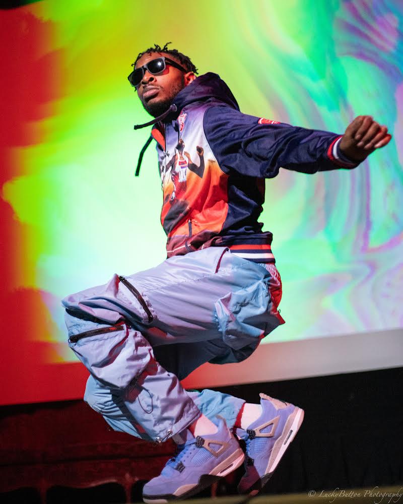 Person jumping with colorful background, wearing sunglasses, a jacket, baggy pants, and sneakers.