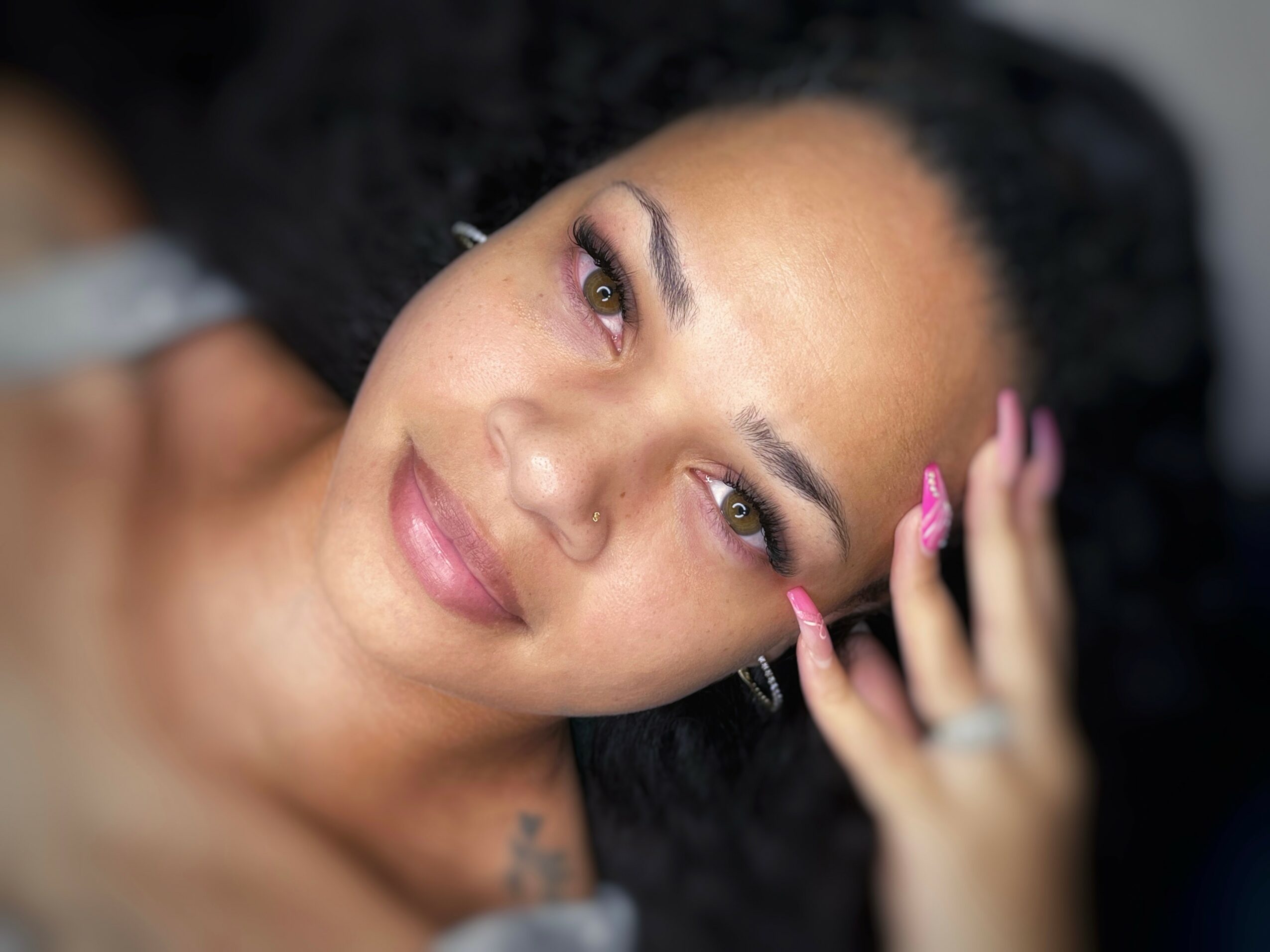 Close-up of a woman with dark curly hair, pink makeup, and pink nails, resting her hand on her face.