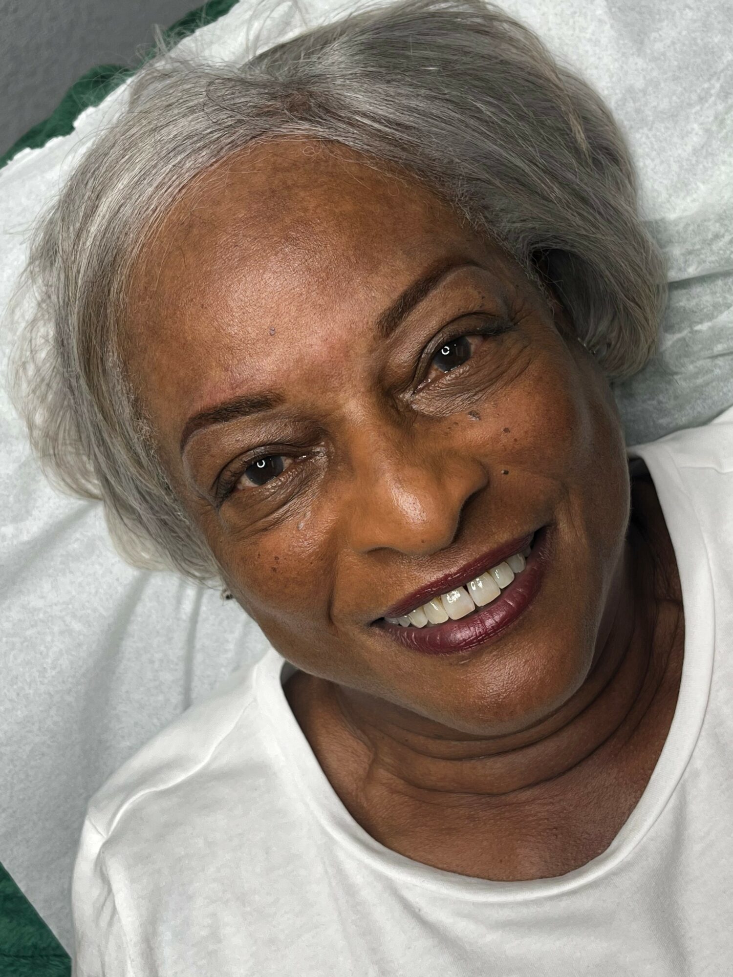 Close-up of smiling elderly woman with gray hair lying on a pillow, wearing a white top.