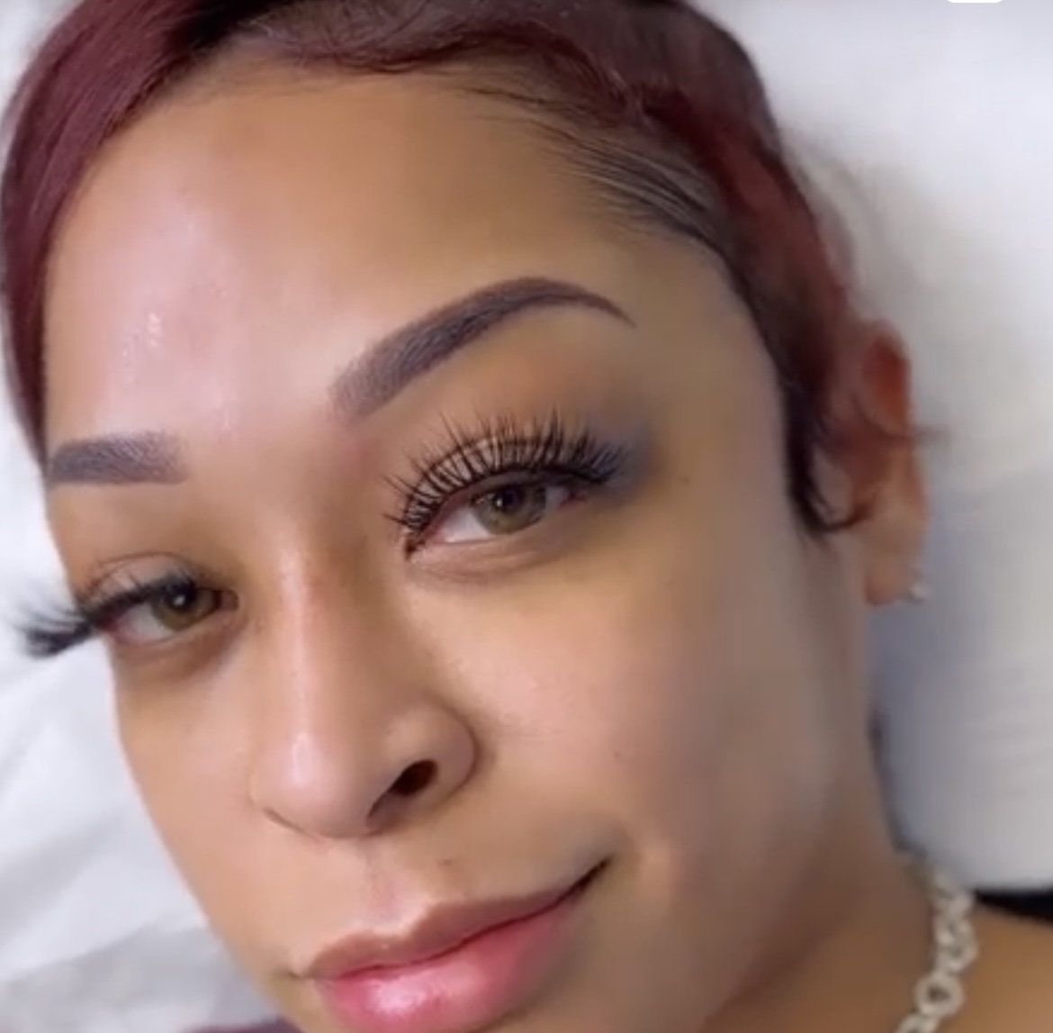 Close-up of a woman's face with makeup, lying down on a pillow, smiling slightly.
