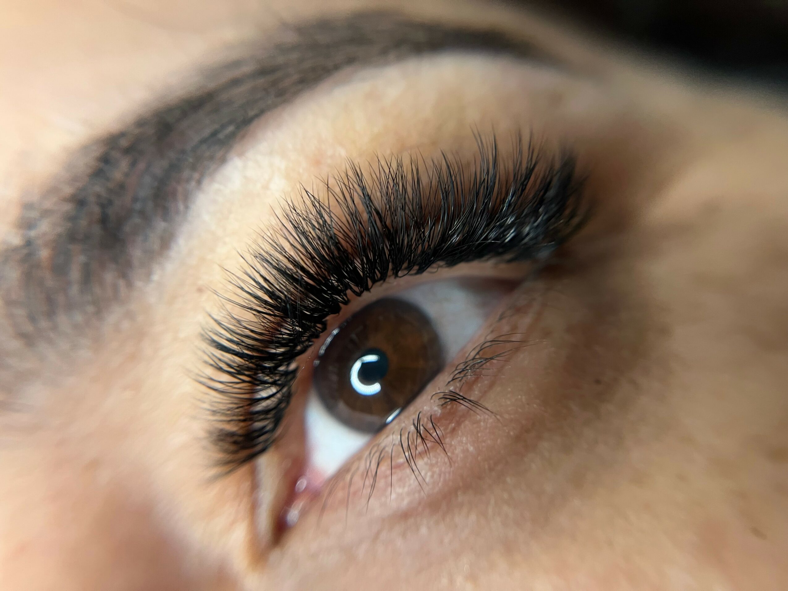 Close-up of a person's eye with long, thick eyelashes and well-defined eyebrow, brown iris, and smooth skin.