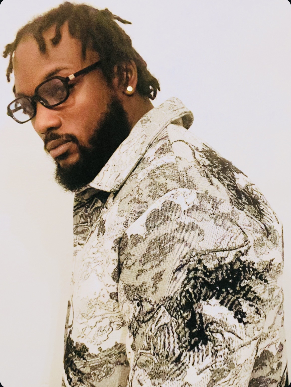 Person with glasses and dreadlocks looking down, wearing a patterned hoodie, against a plain background.