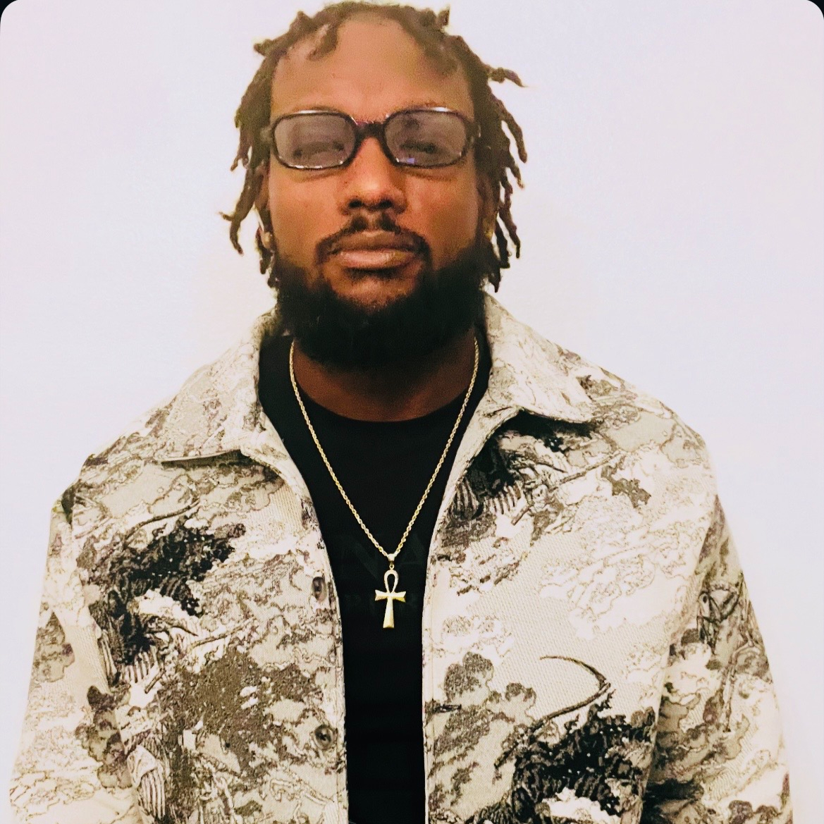 Man with glasses, dreadlocks, beard, wearing a patterned jacket and an ankh necklace, facing forward.