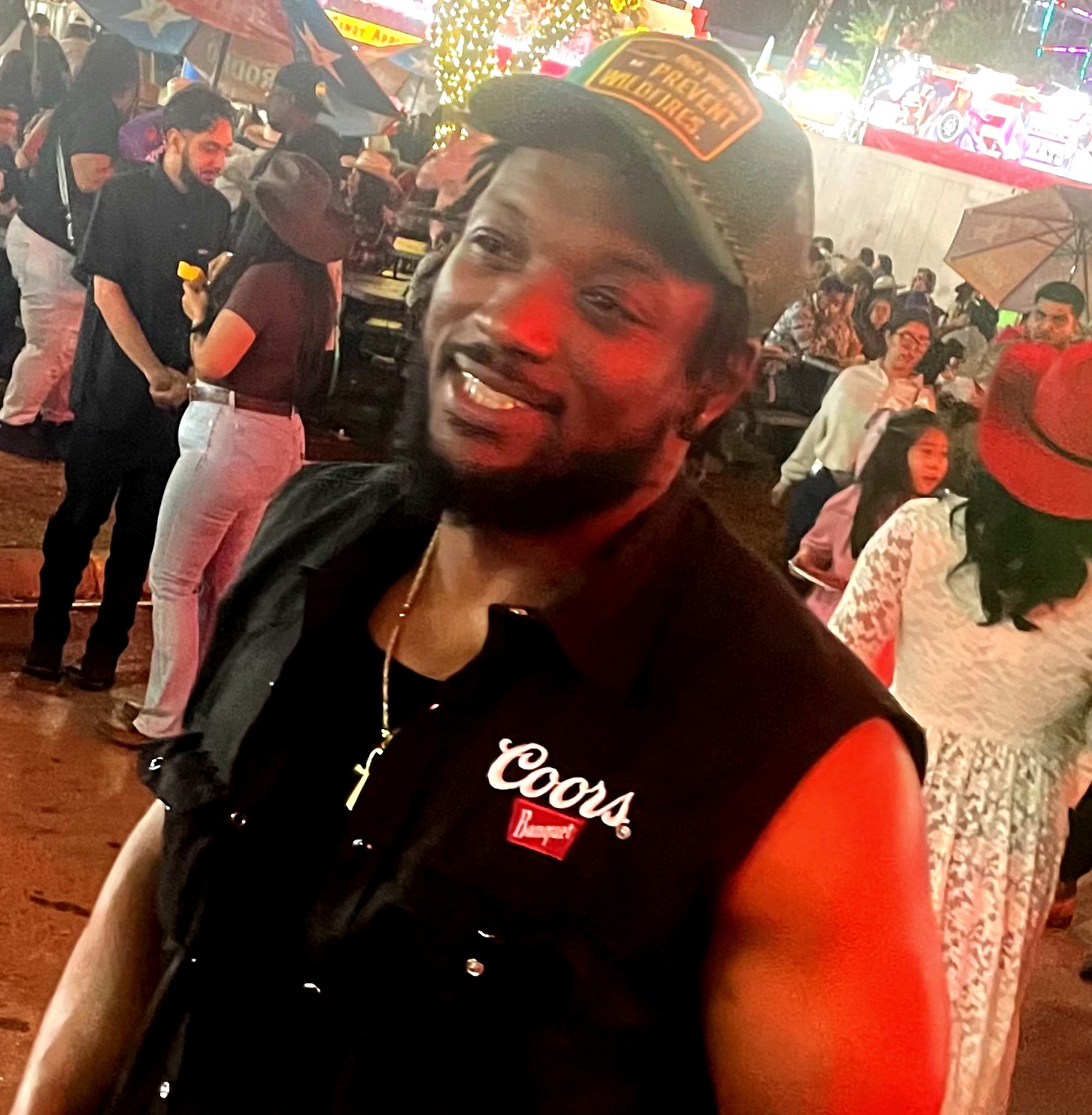 Man smiling at a crowded outdoor event at night, wearing a black vest with patches and a cap, surrounded by people.