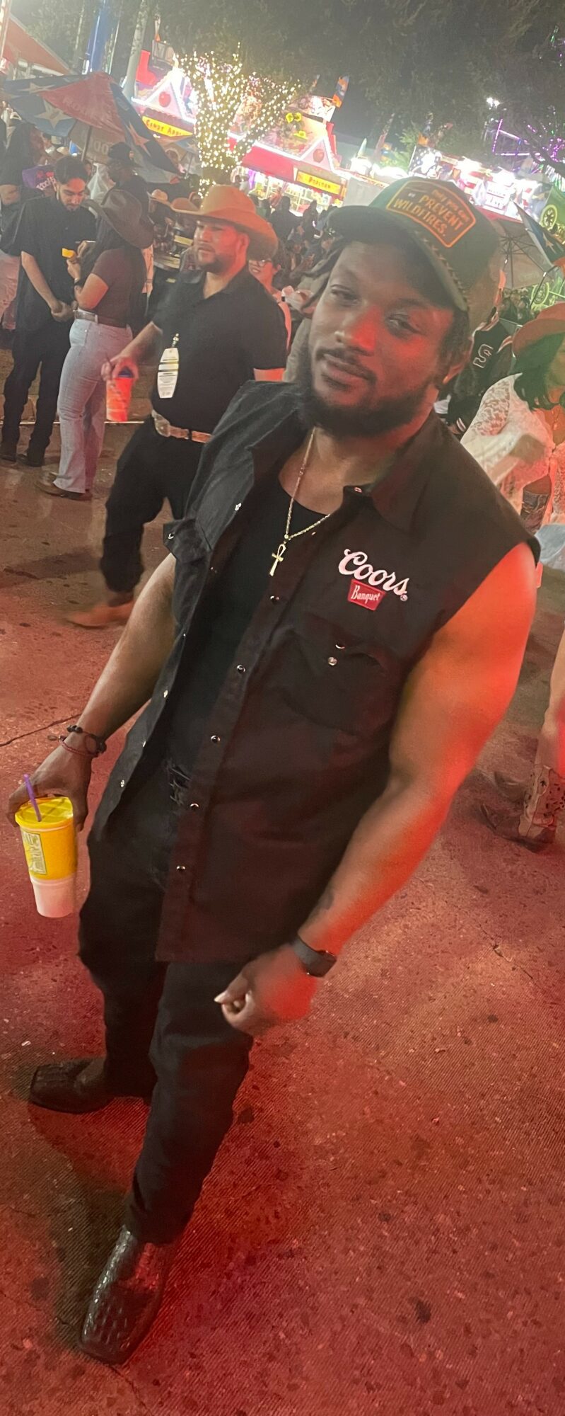 Man with beard wearing a black vest and cap, holding a yellow drink, at an outdoor event at night.