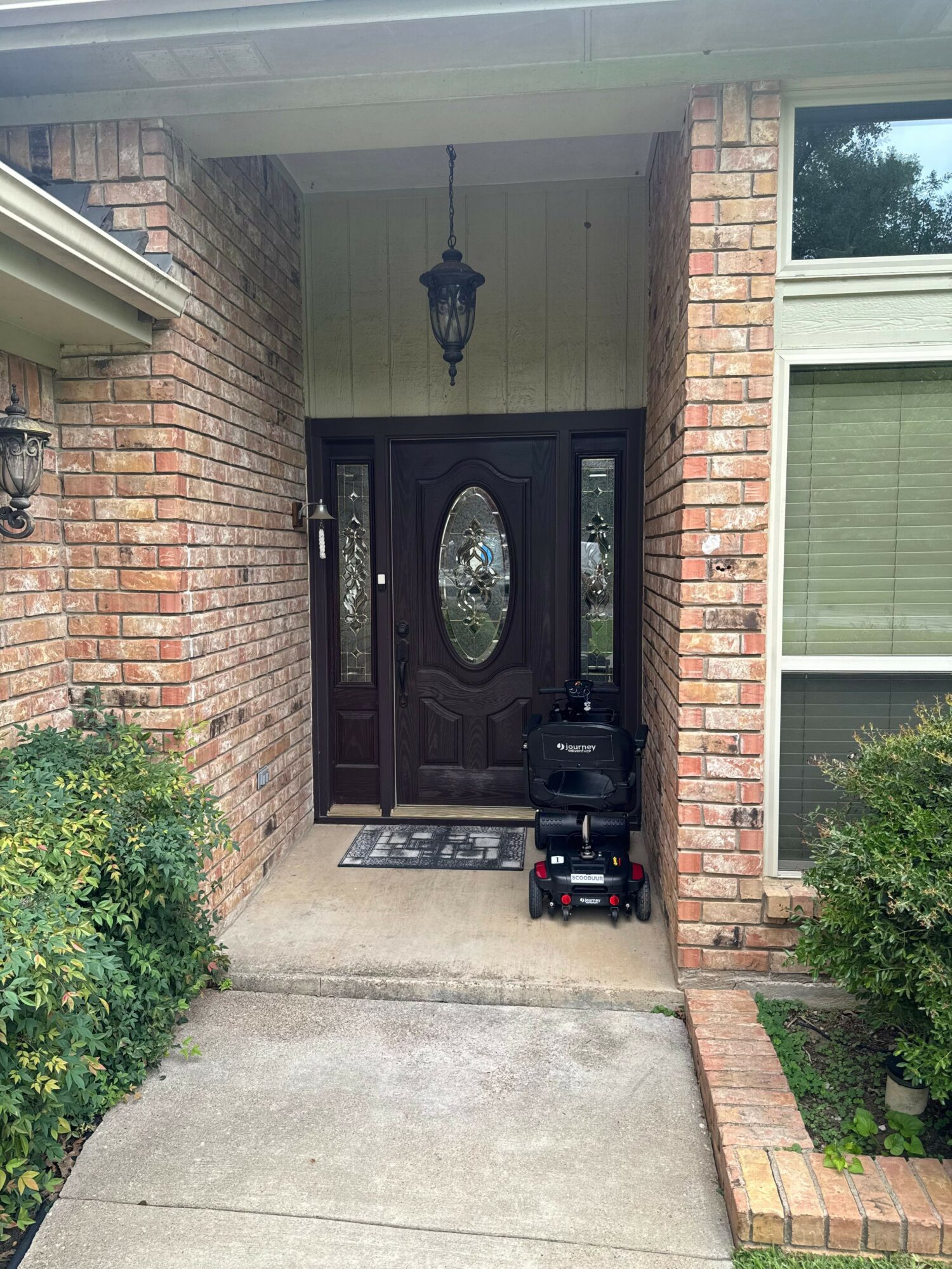 Front door with brick pillars, black door with oval glass, outdoor light, plants, and a scooter on the porch.