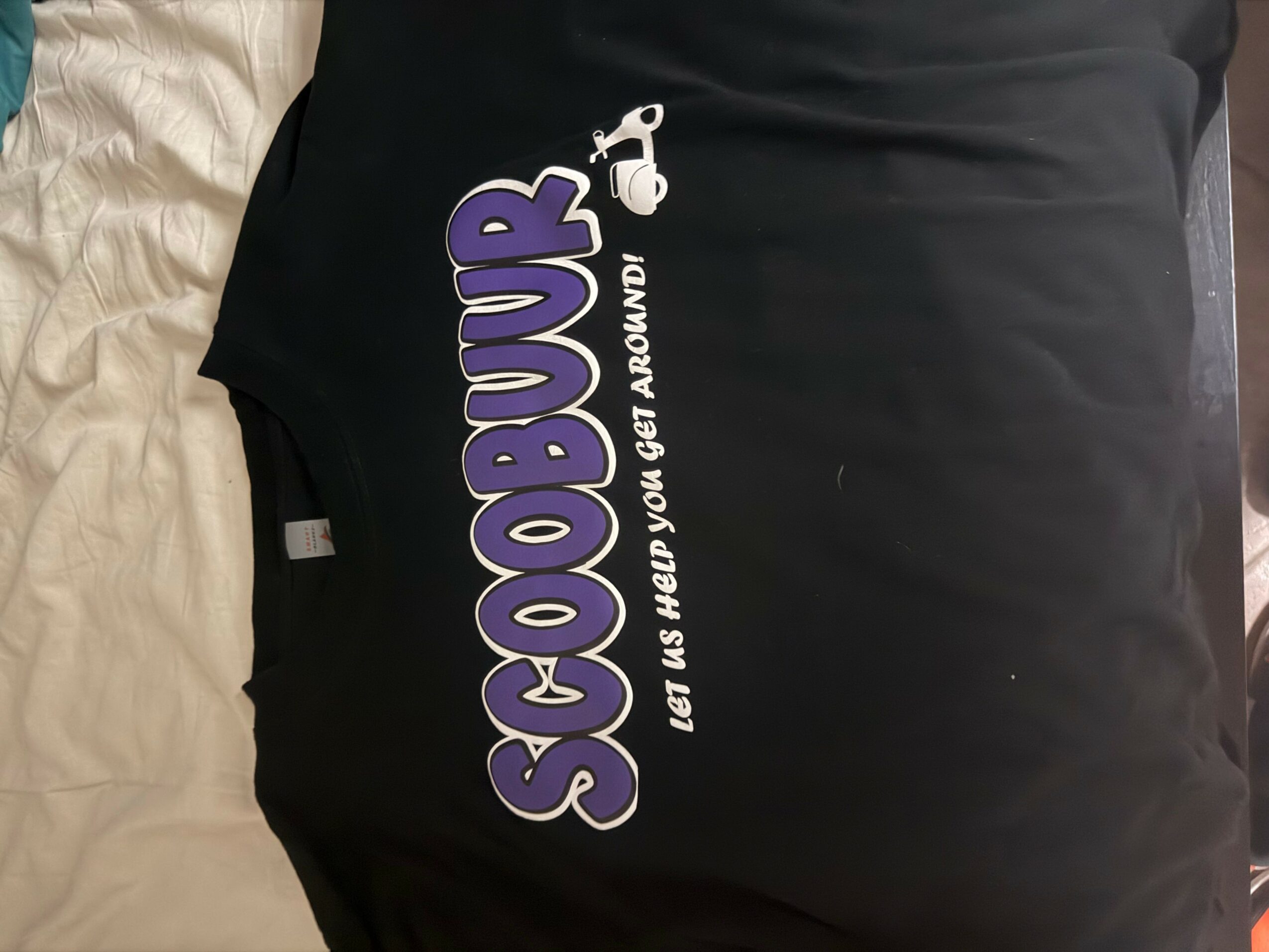 Black shirt with purple and white text and a small graphic, displayed on a bed.