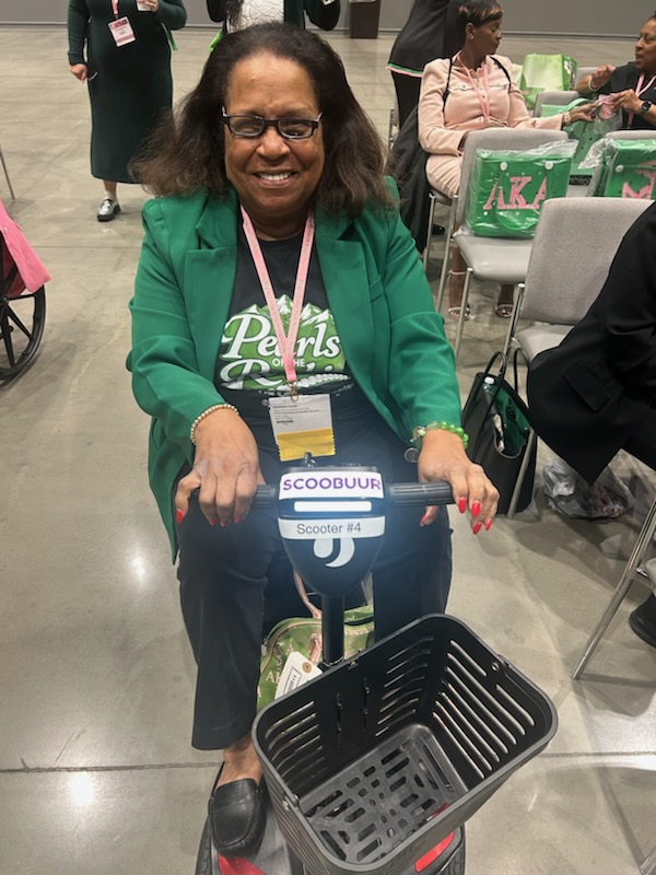 Smiling woman with glasses and curly hair, wearing a green blazer, standing behind a scooter with a basket, indoors.