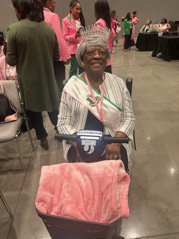 Elderly woman wearing a crown and sash, sitting in a wheelchair with a pink blanket, at an indoor event with people in the background.