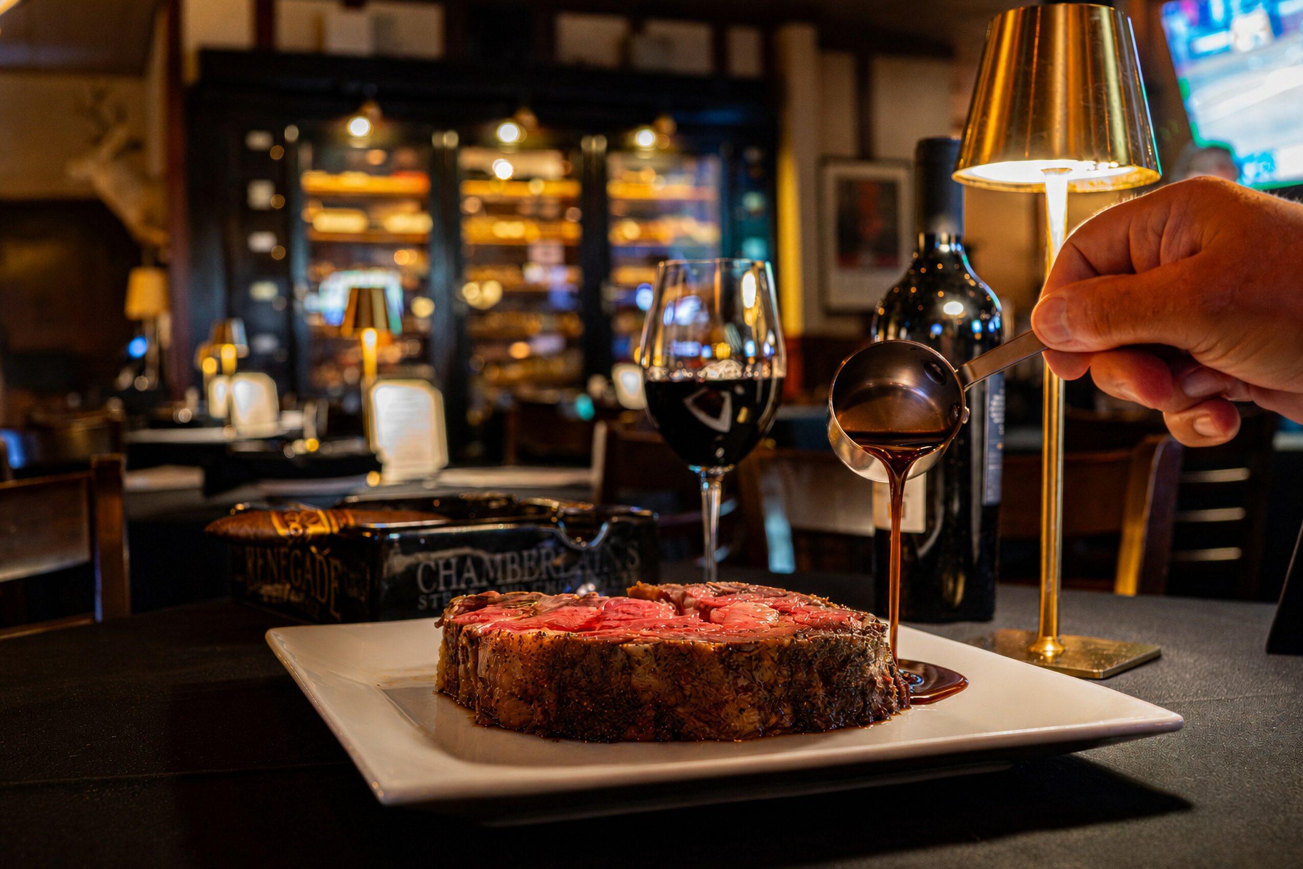 A steak on a white plate with a glass of red wine, a hand holding a spoon, and a warm-lit bar background.