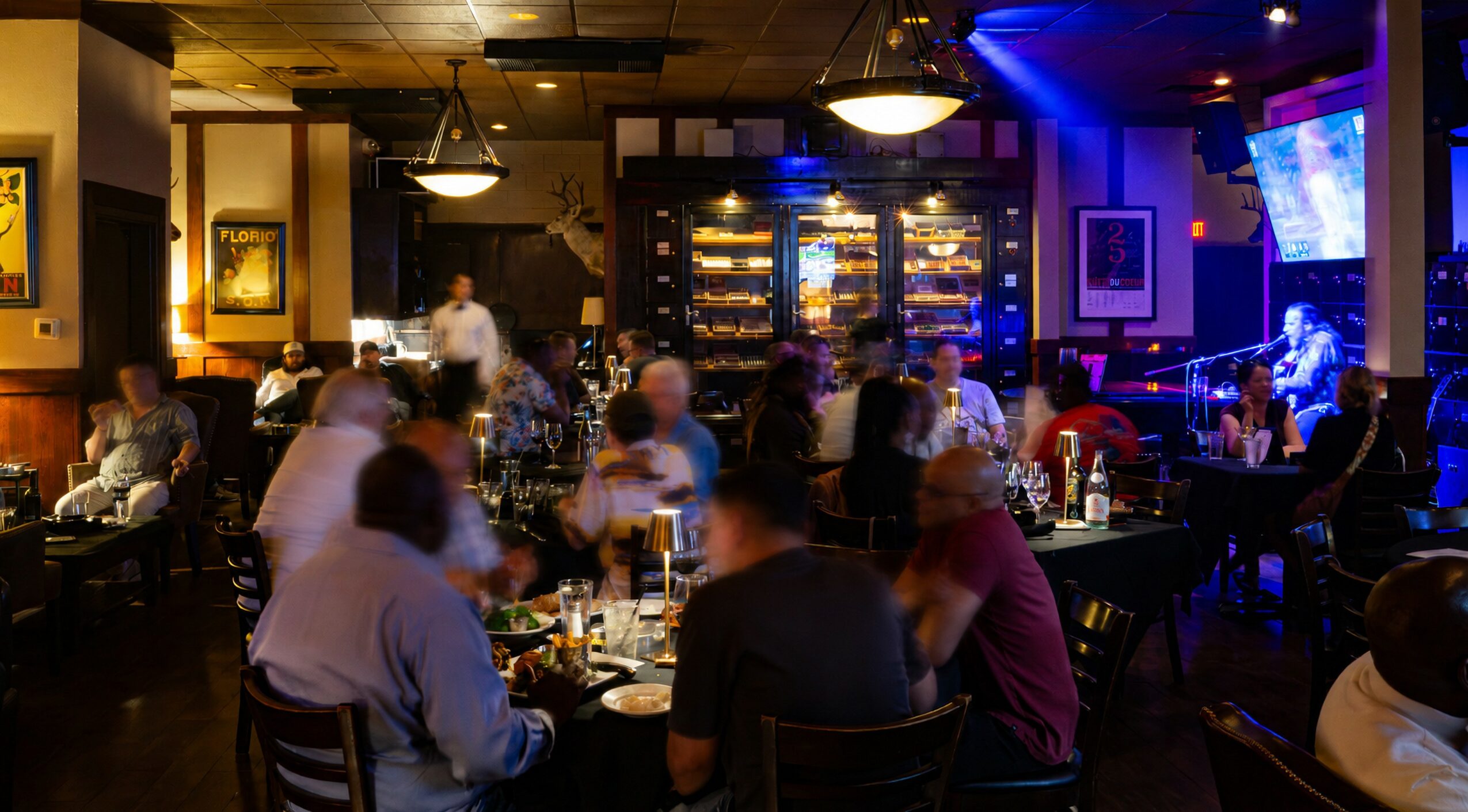 People dining and socializing in a dimly lit restaurant with a bar and television screens, warm lighting, and a cozy atmosphere.