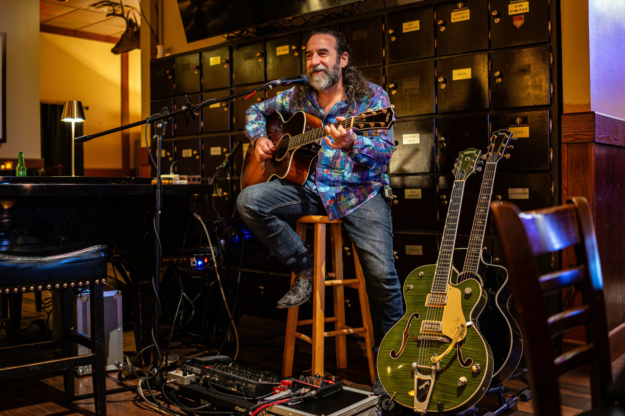Man playing guitar on a stool with another guitar on the floor beside him, in a room with musical equipment.