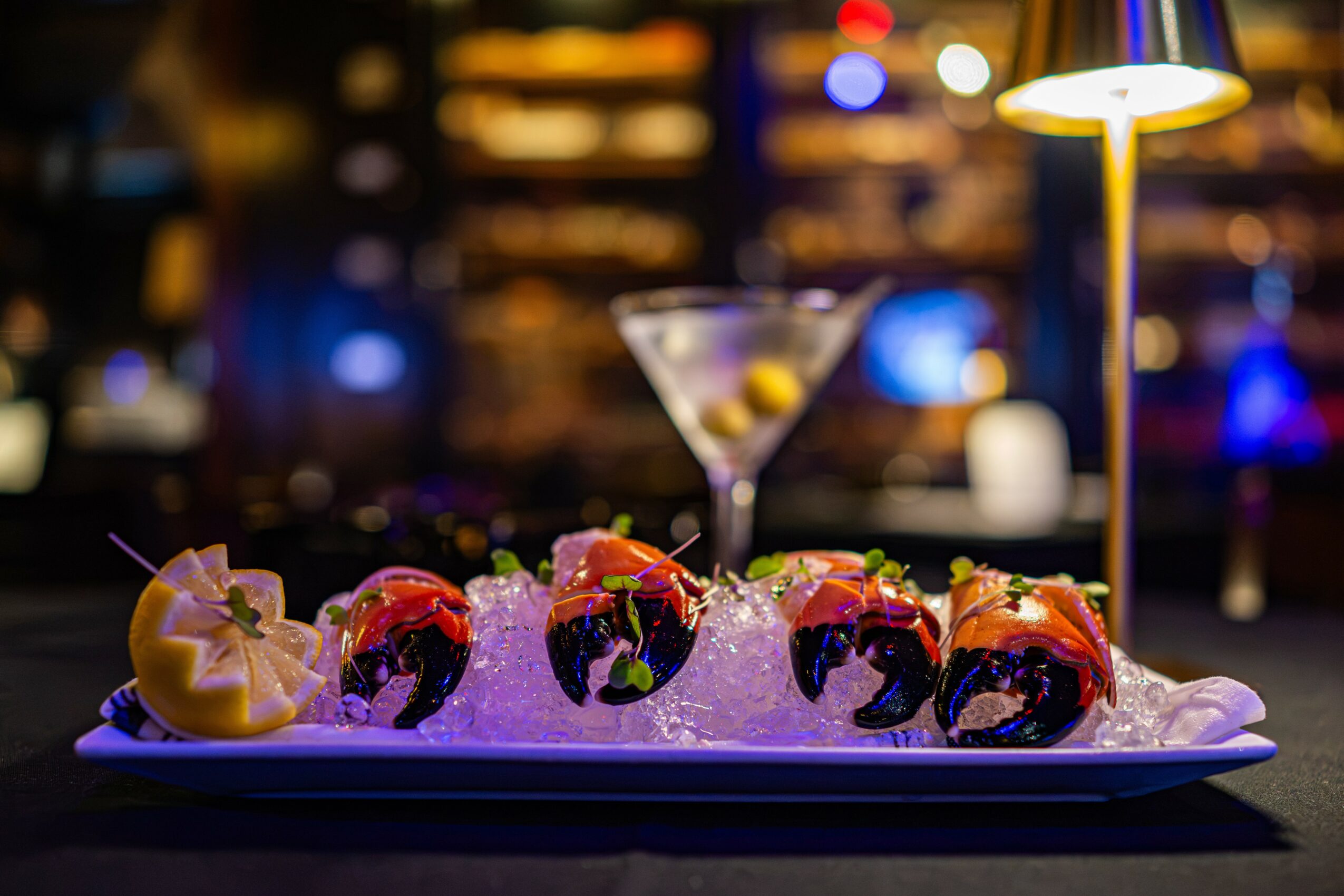 Sushi rolls on a plate with ice, a martini glass, and a lamp in a dimly lit bar setting.