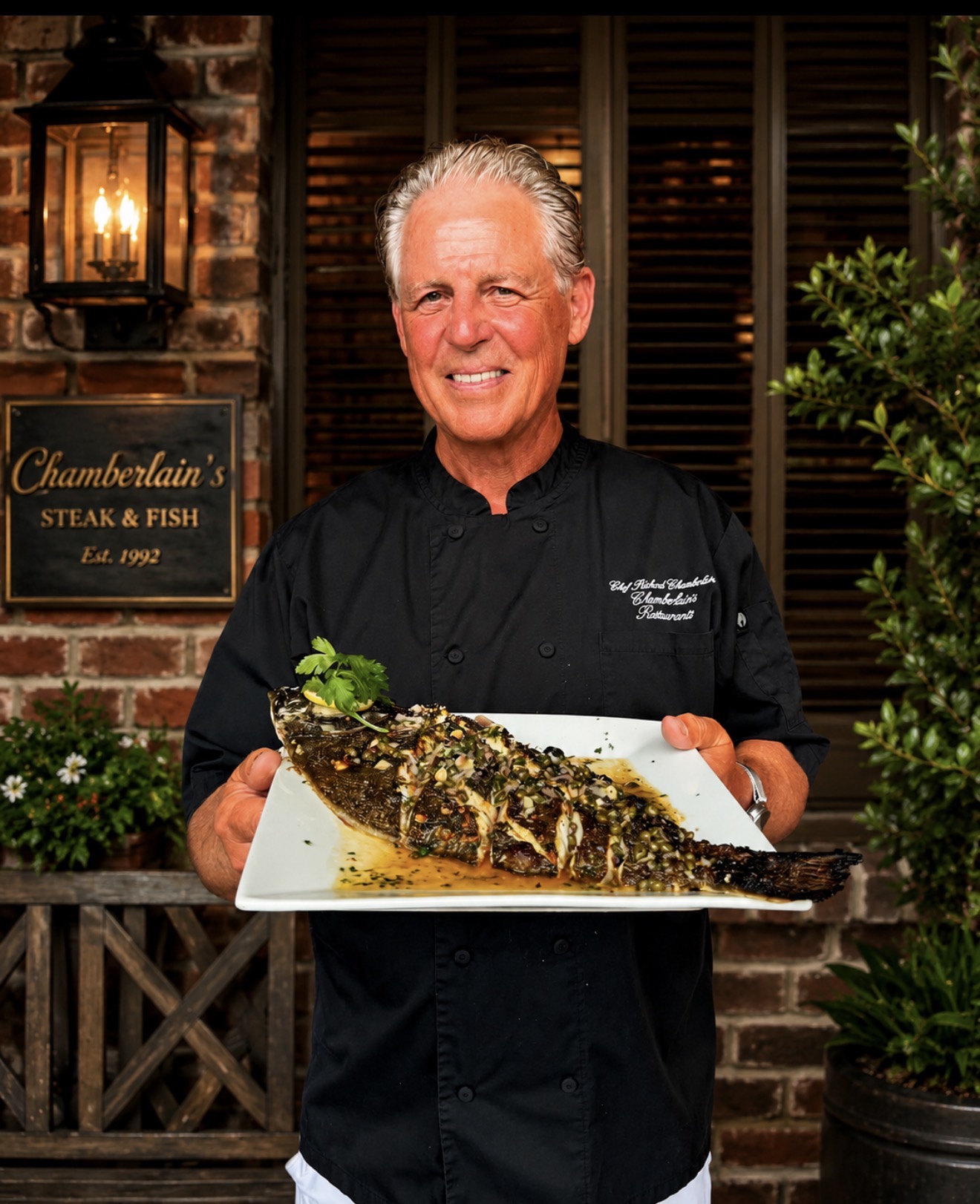 Chef holding a large plated fish dish outdoors, smiling, with brick wall, lantern, and greenery in background.
