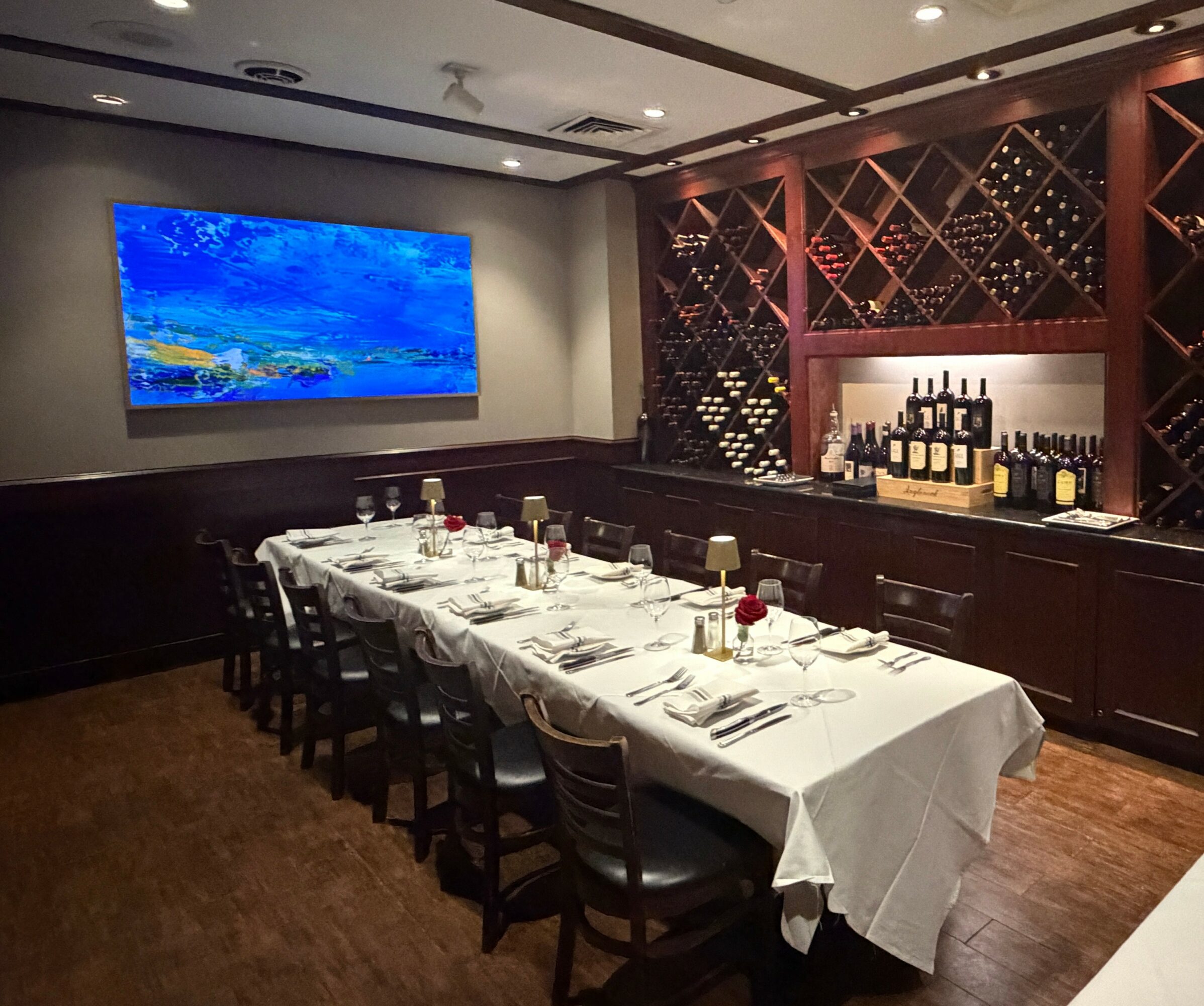 Dining room with a long table set with white tablecloths, wine bottles on shelves, and a large landscape painting on the wall.