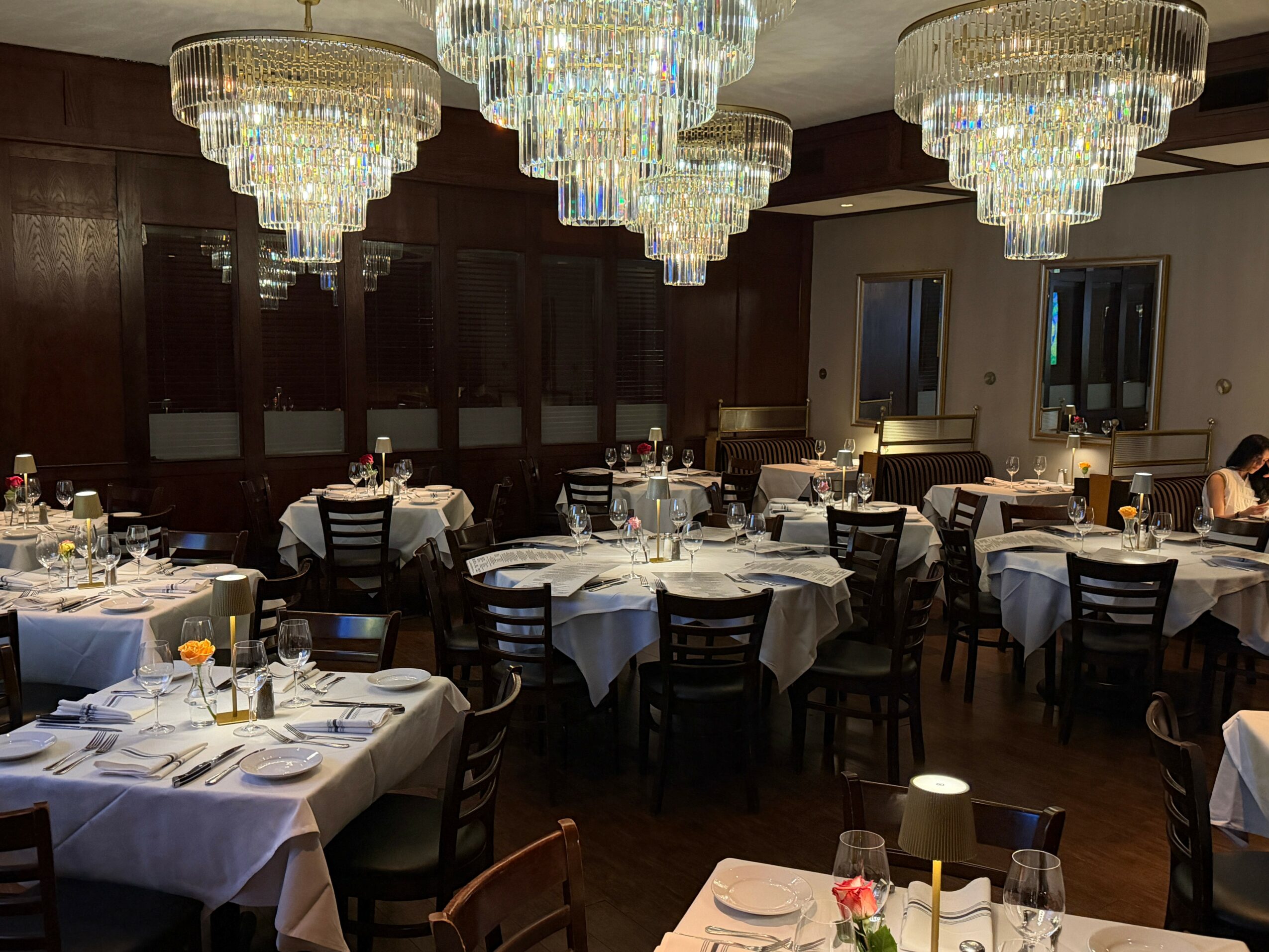Elegant dining room with chandeliers, round tables with white tablecloths, set with plates, glasses, and silverware, dark wood walls.