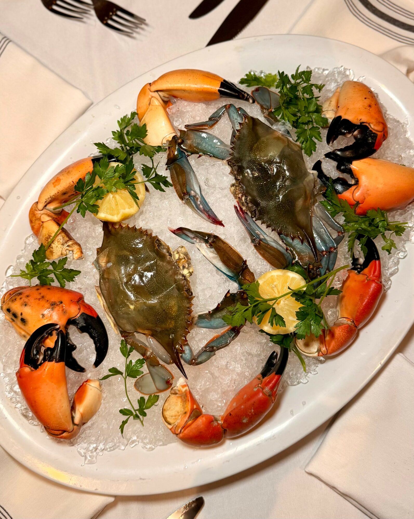 Fresh crabs on a bed of ice garnished with lemon wedges, parsley, and sliced vegetables on a white platter.