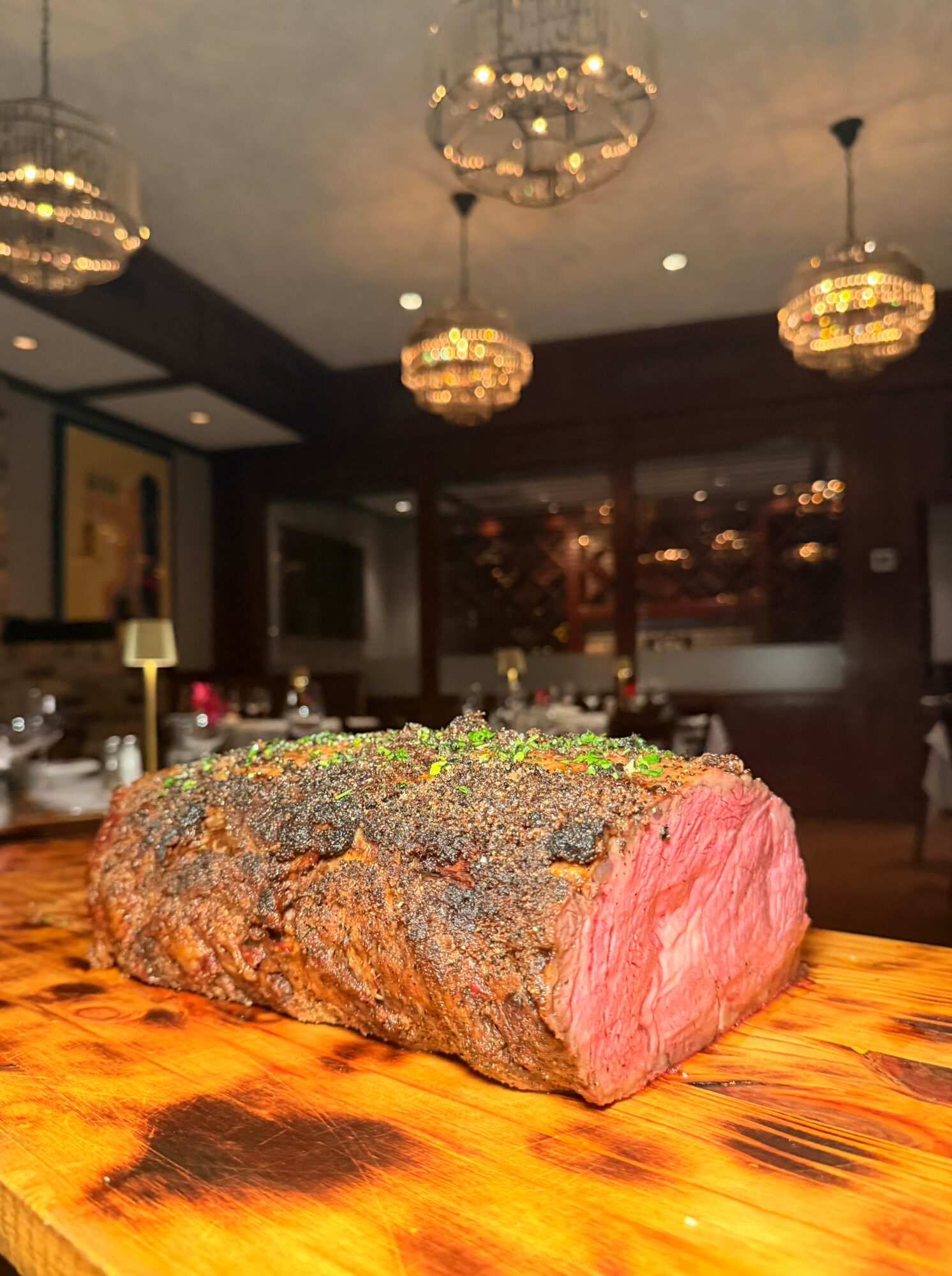 A large piece of cooked meat with a pink interior and a dark, crispy exterior on a wooden table in a dimly lit room.