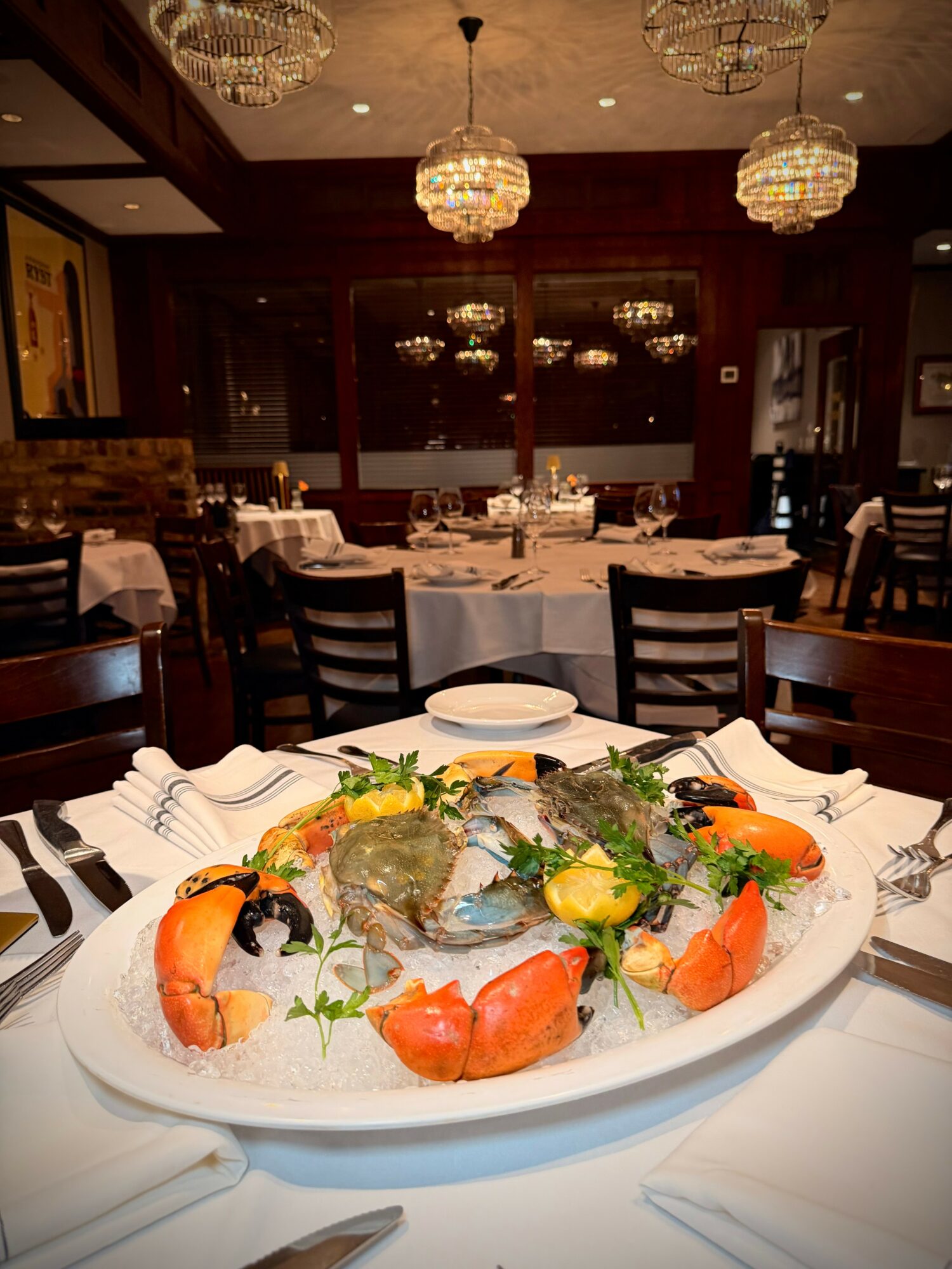 Elegant restaurant dining room with round tables, chandeliers, and a large seafood platter with fish, vegetables, and lemon slices.