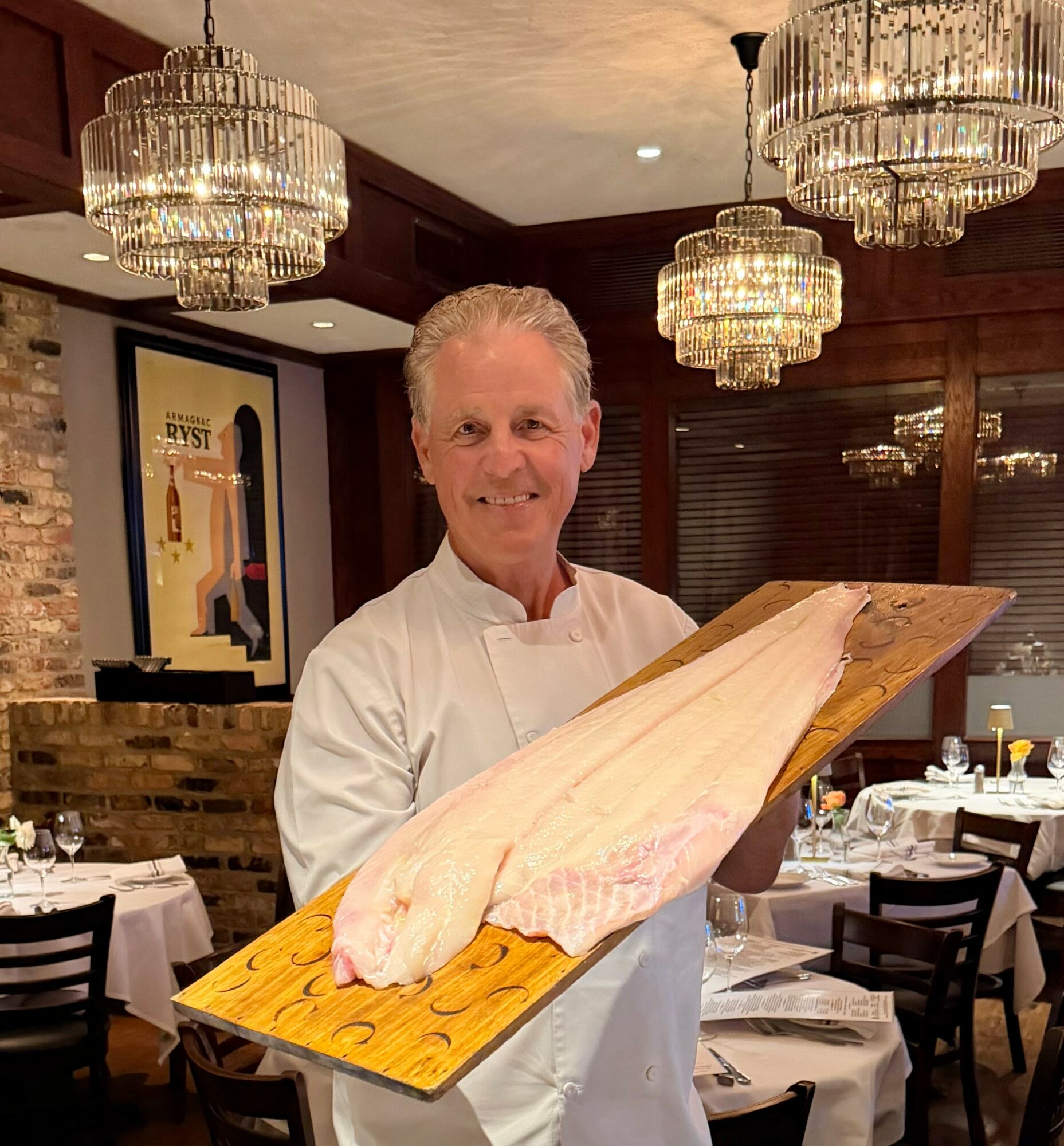 Chef holding a large fish on a wooden board in a restaurant with chandeliers and tables.