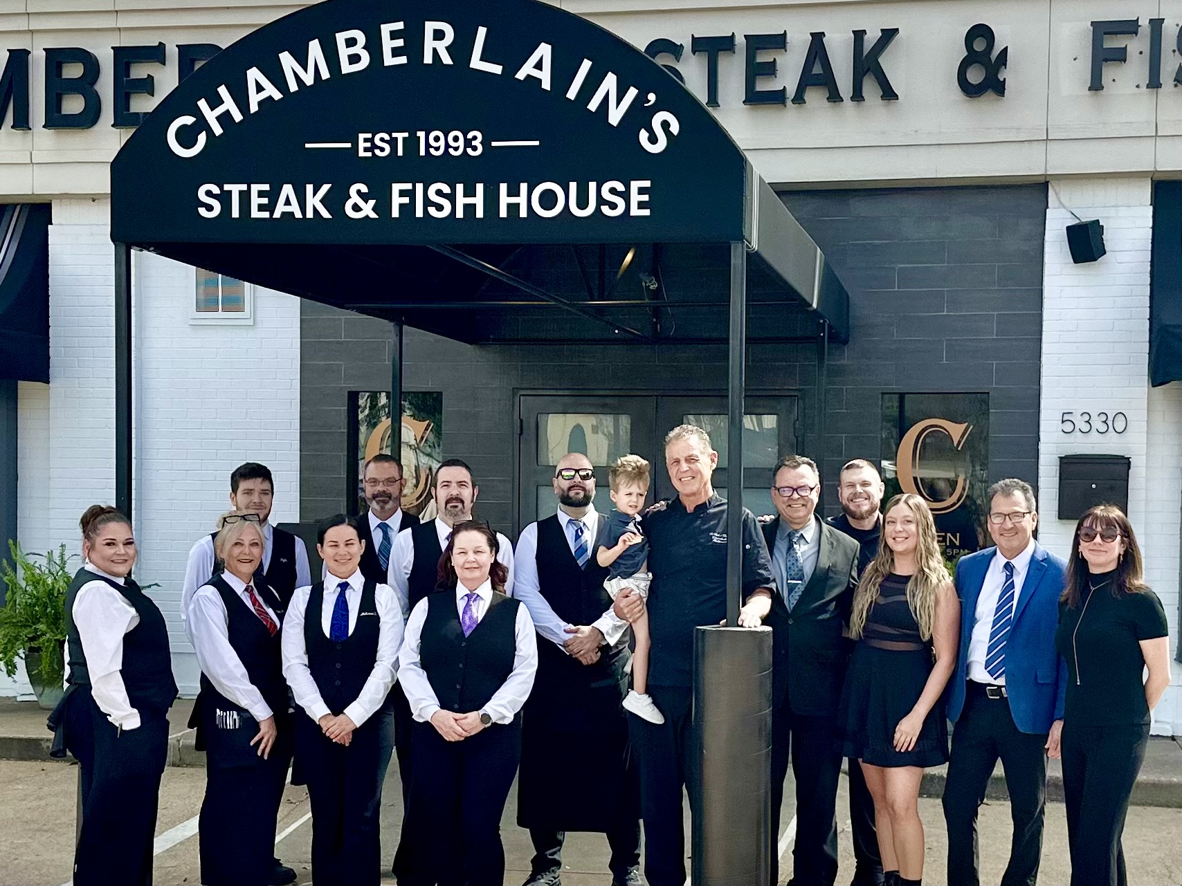 Group of people standing outside a restaurant with a sign reading 'Chambertain's Steak & Fish House,' in front of a building with a gray facade.