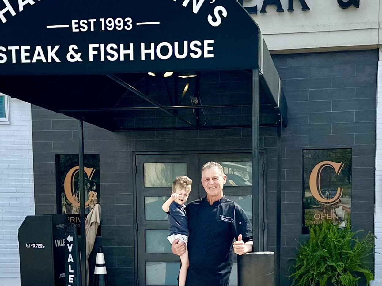 Man holding a child in front of a restaurant entrance, smiling and giving a thumbs-up, with a sign reading 'EST 1993' and 'STEAK & FISH HOUSE'.