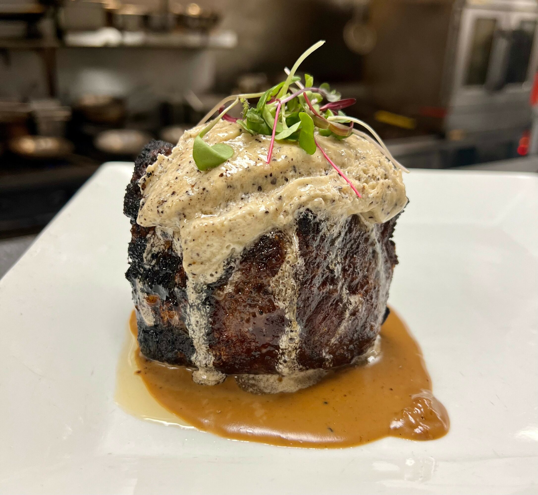 A cooked meat dish topped with sauce and microgreens, served on a white plate with sauce underneath, in a restaurant setting.