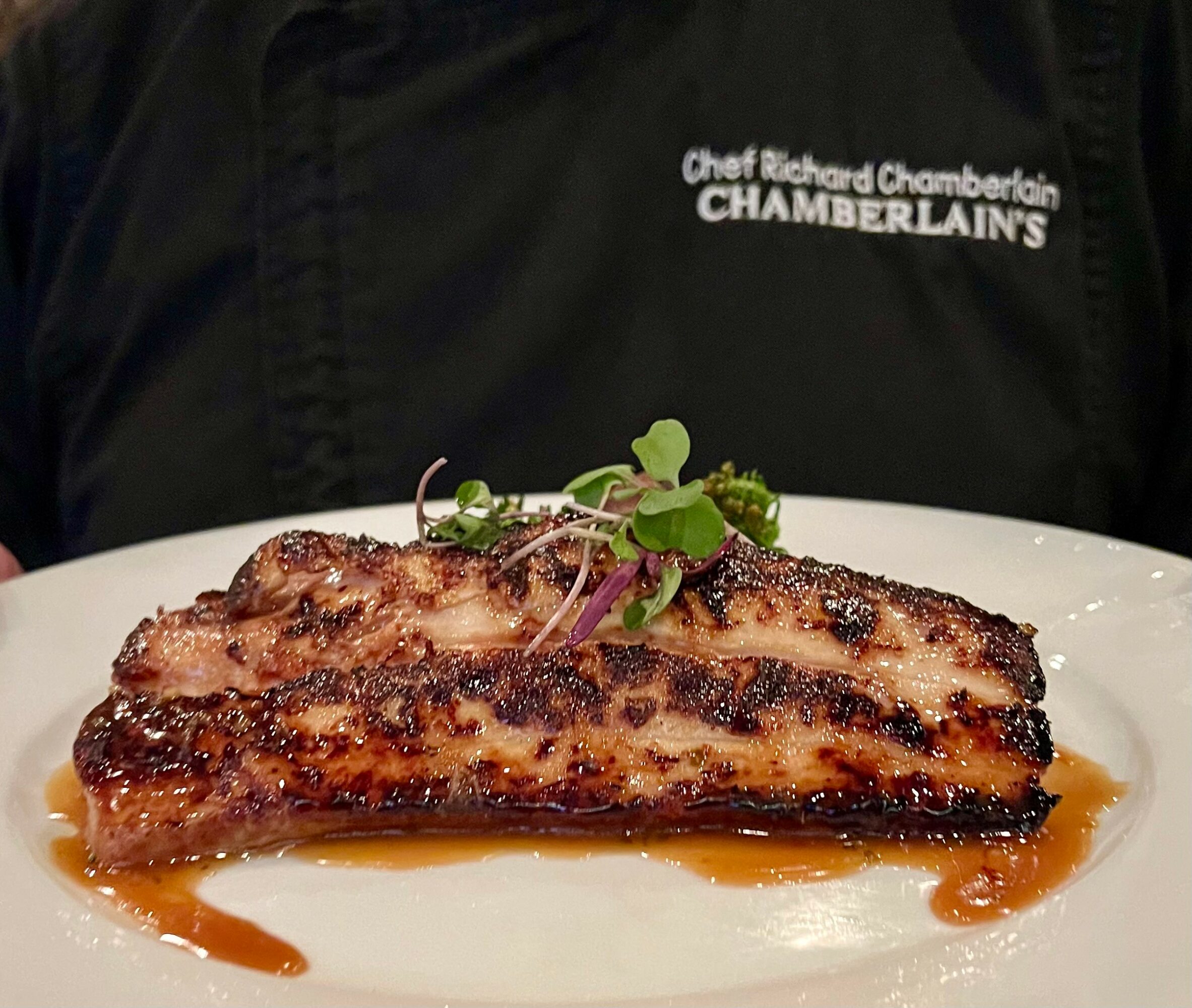 A grilled meat steak garnished with microgreens on a white plate, with sauce underneath, held by a person in a black chef's jacket.
