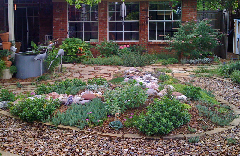 Garden with plants, rocks, and a brick building with large windows in the background.