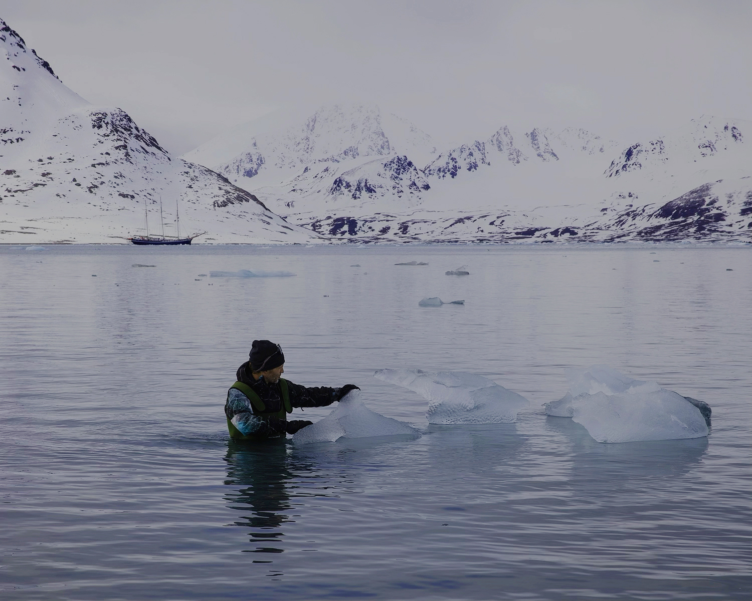 Person in water near ice chunks with snow-covered mountains in background.