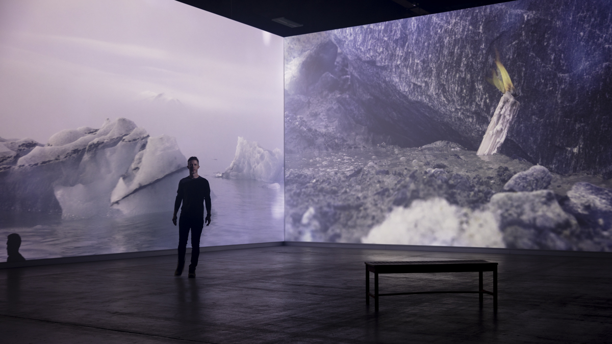 Person standing in front of large digital screens displaying icy landscape and water scene, with a bench nearby.