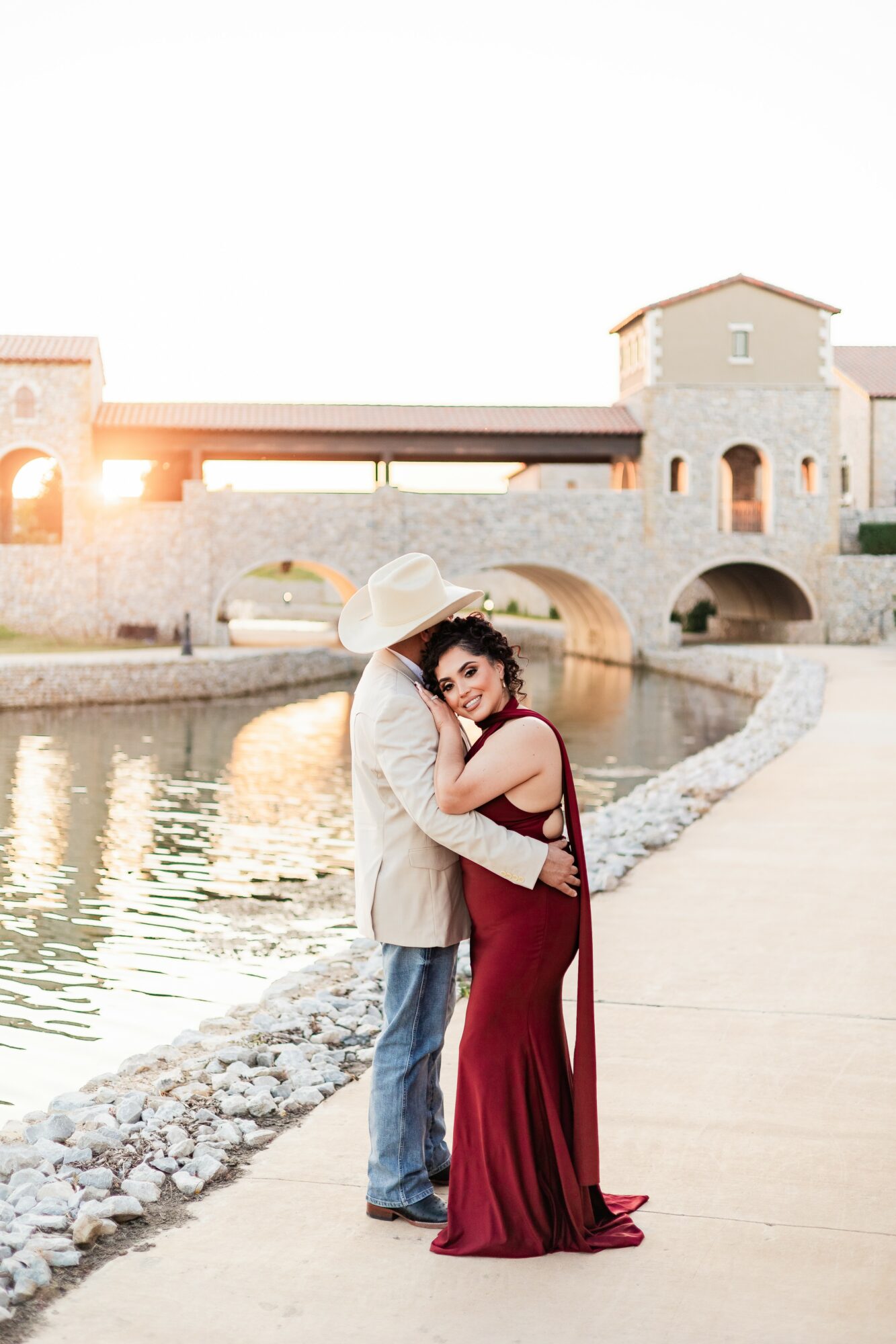 A couple hugging by a river with historic buildings and a bridge in the background during sunset.