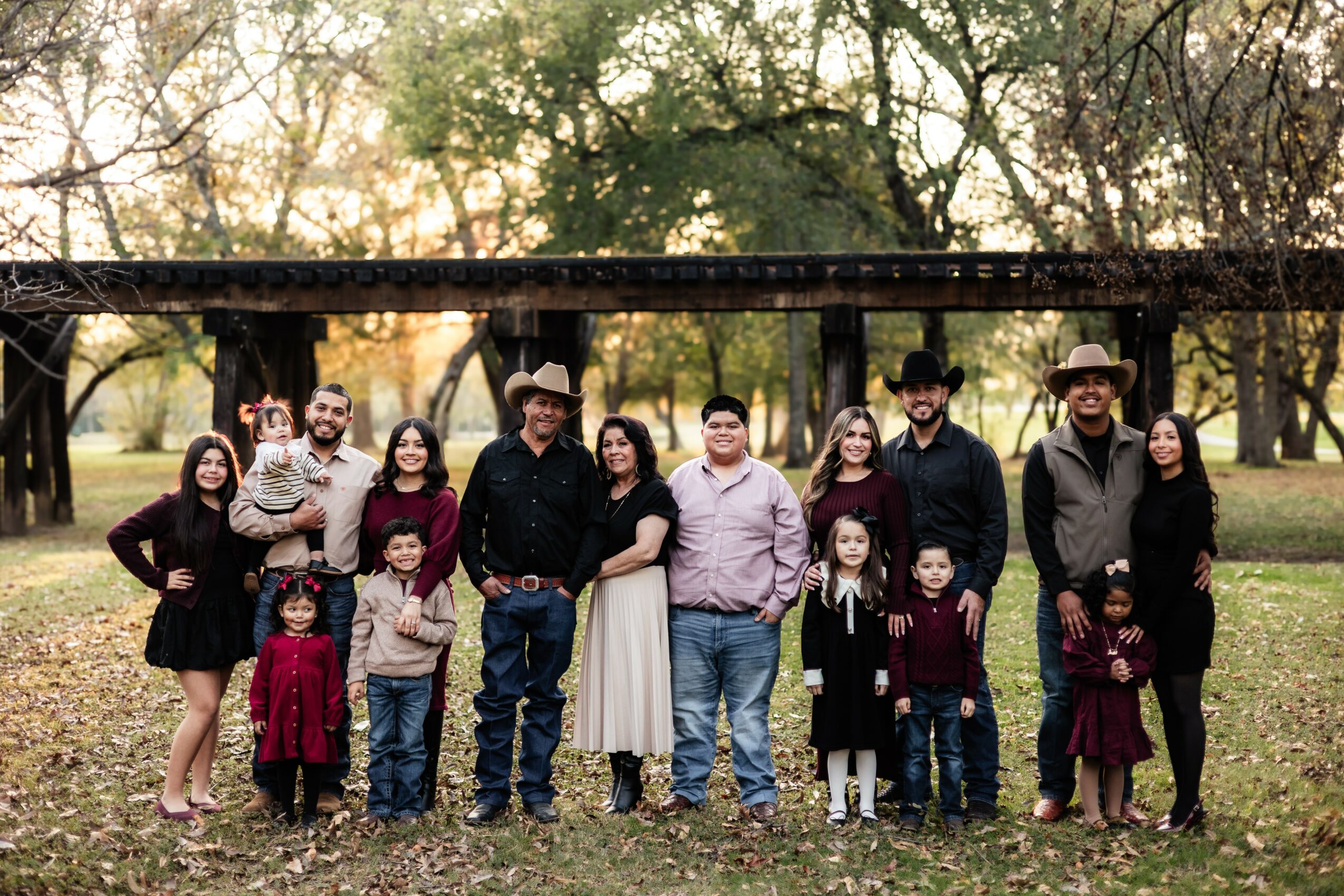 Group of people standing outdoors in a park with trees, some children, and adults smiling, dressed casually, during daytime.