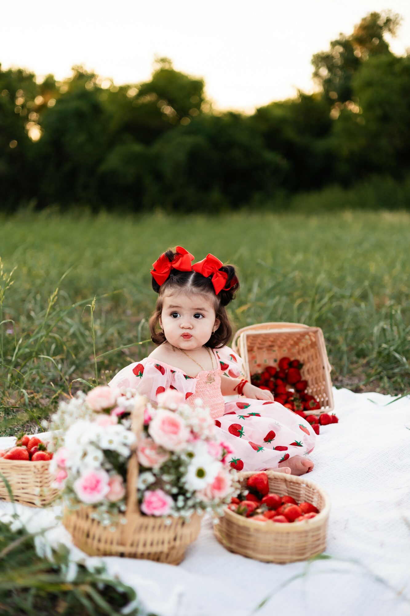 Young girl in a red and white dress with strawberries, sitting on a white cloth outdoors with baskets of strawberries and flowers.