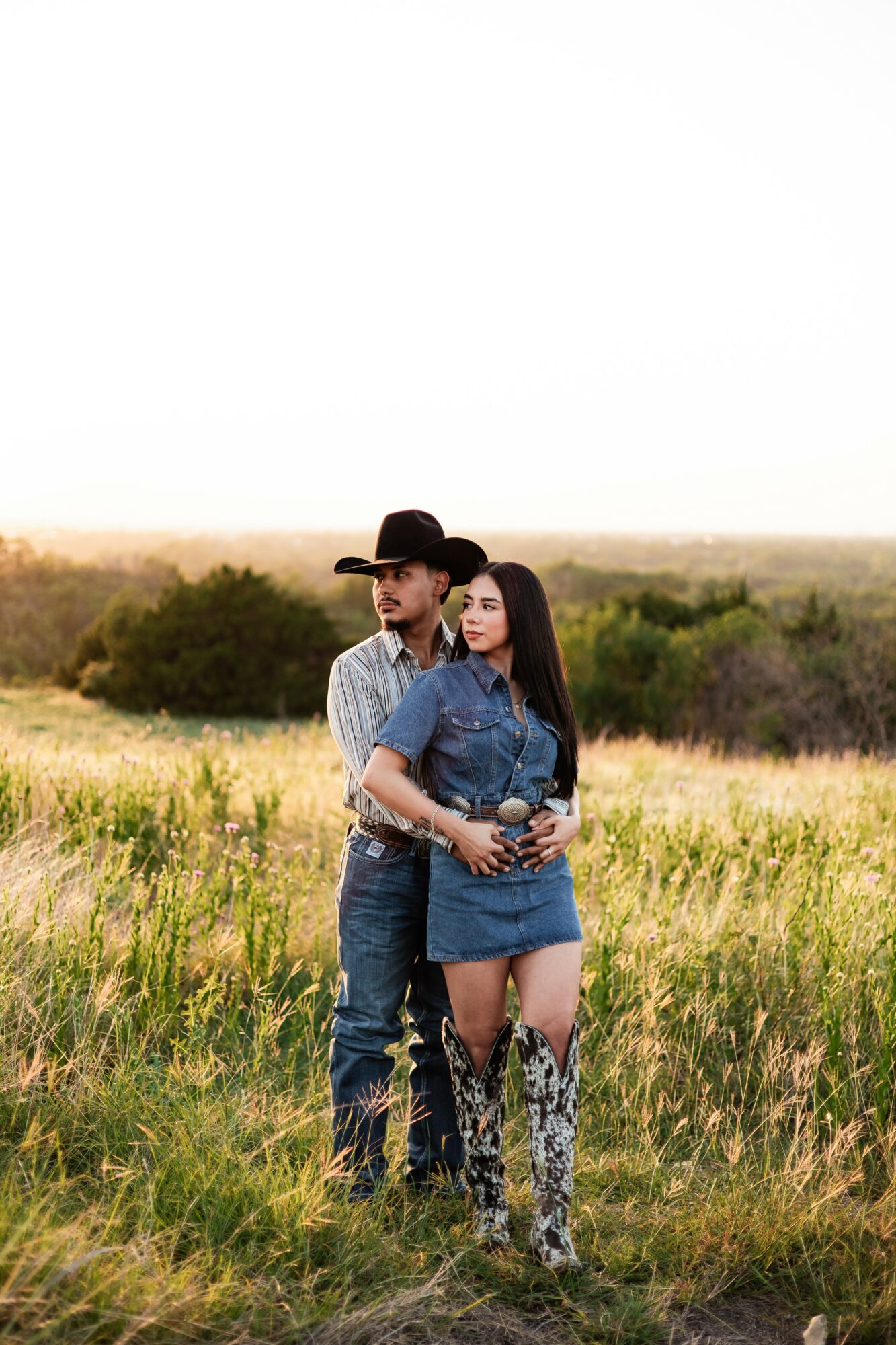 A couple stands in a grassy field with bushes and a horizon in the background, embracing each other.