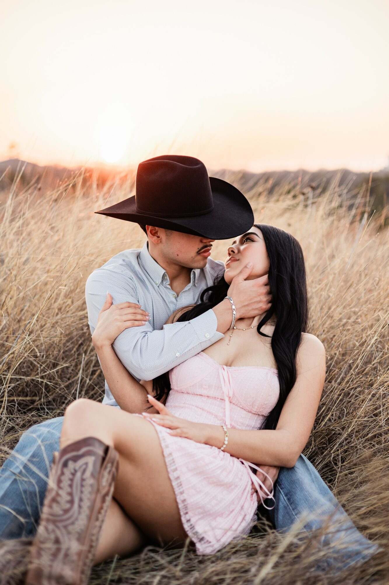 A couple sitting in a field at sunset, the man wearing a hat and the woman in a pink dress, embracing.