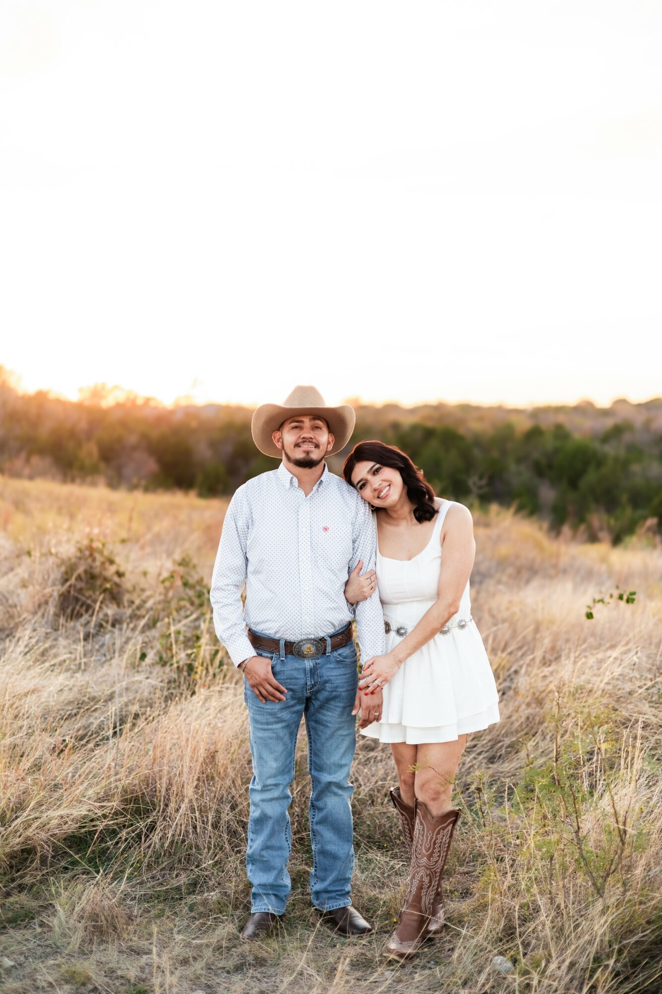 A man and woman stand together in a grassy field, smiling, with trees and a clear sky in the background.