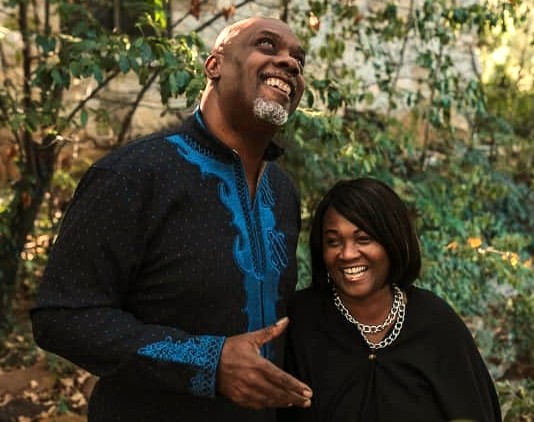 Two smiling people outdoors, one taller man with a beard and a woman with short hair, surrounded by greenery.