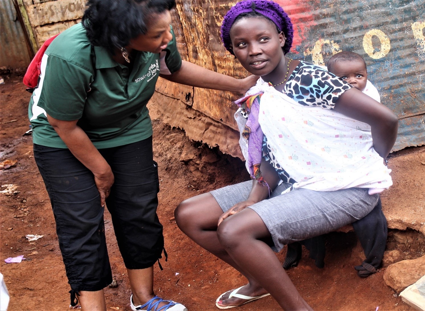 Woman in green shirt interacts with a seated girl wearing a purple headscarf and white top, with a boy in the background.
