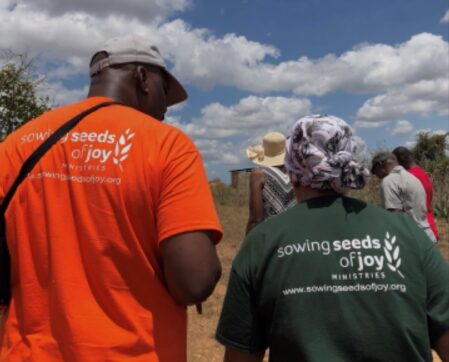 Two people wearing shirts with 'sowing seeds of joy' text, outdoors with a group and cloudy sky.