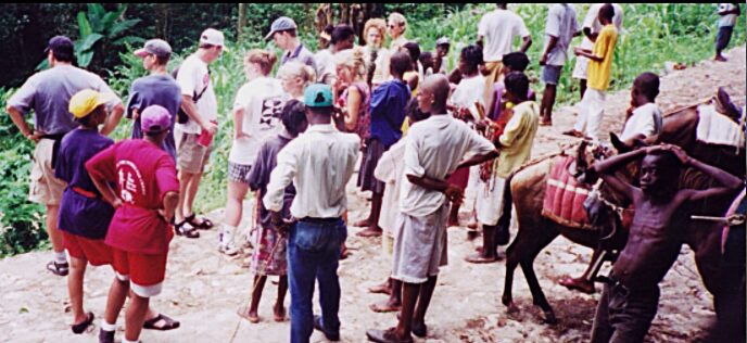Group of people gathered outdoors, some with animals, in a rural setting with trees and dirt ground.