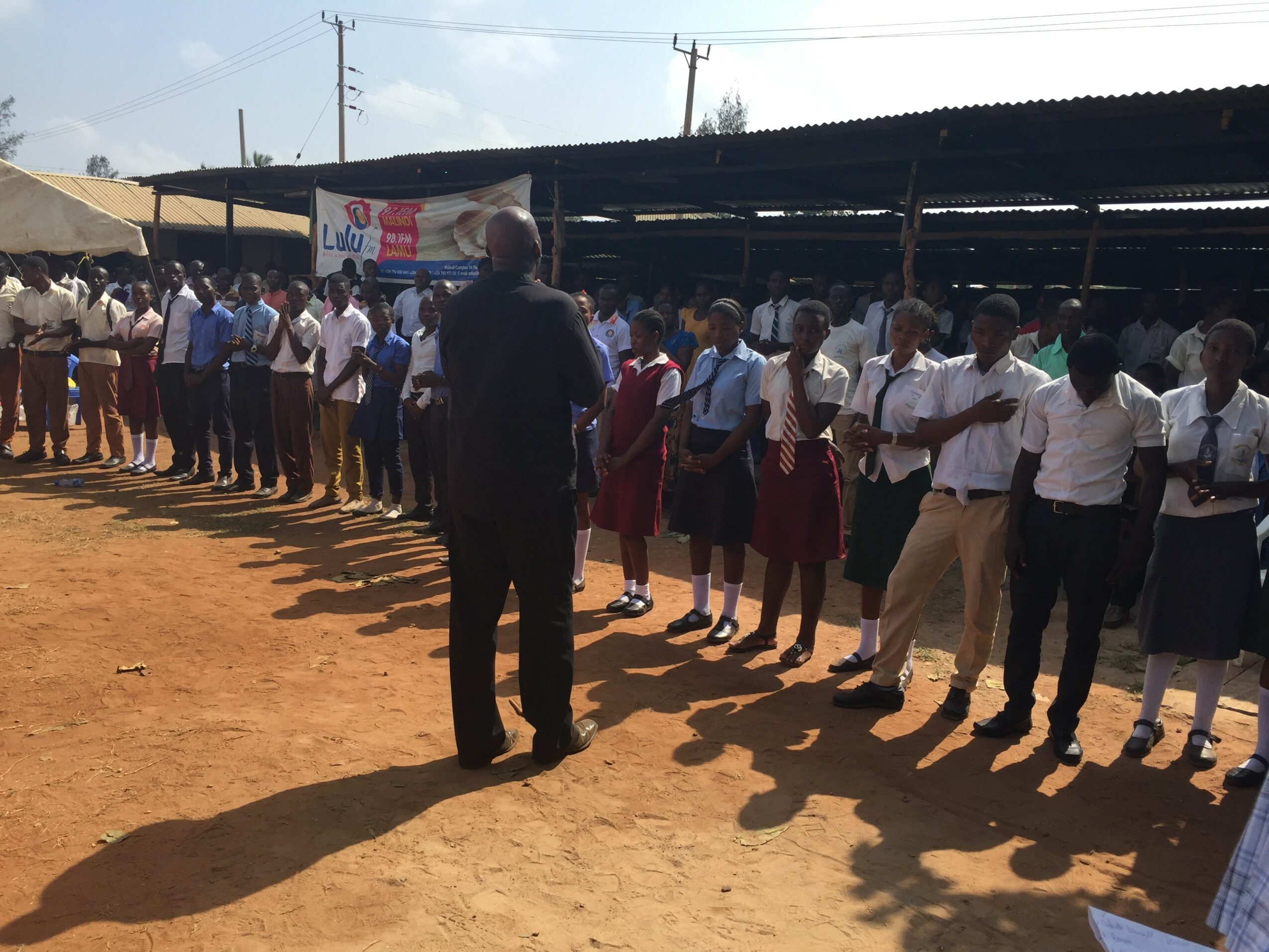 Group of students standing in line outdoors, with a person speaking to them, under a shaded structure, on a dirt ground.