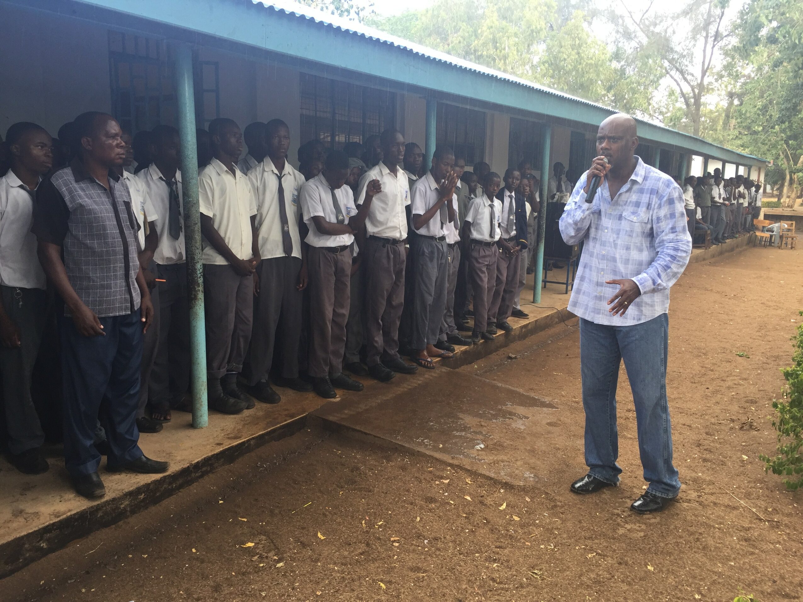 A group of students in school uniforms standing outside a building, listening to a man speaking with a microphone.
