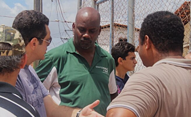 Group of people outdoors, engaged in conversation, with a chain-link fence and structures in the background.