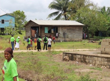 Group of children and adults gather outside a wooden house with a metal roof, surrounded by greenery and palm trees.