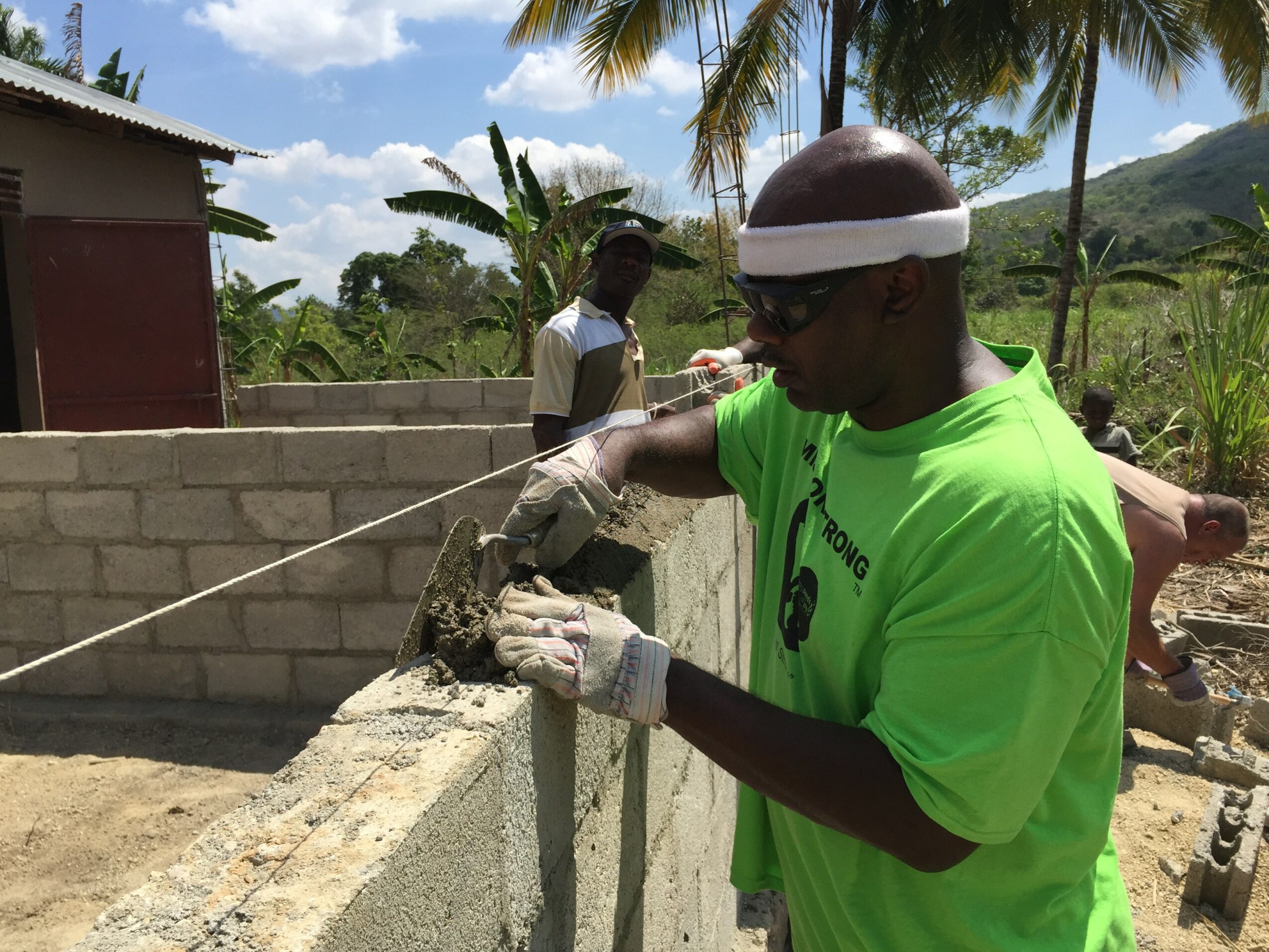 Man in bright green shirt and white headband working on a concrete wall outdoors, with tropical trees and mountains in background.