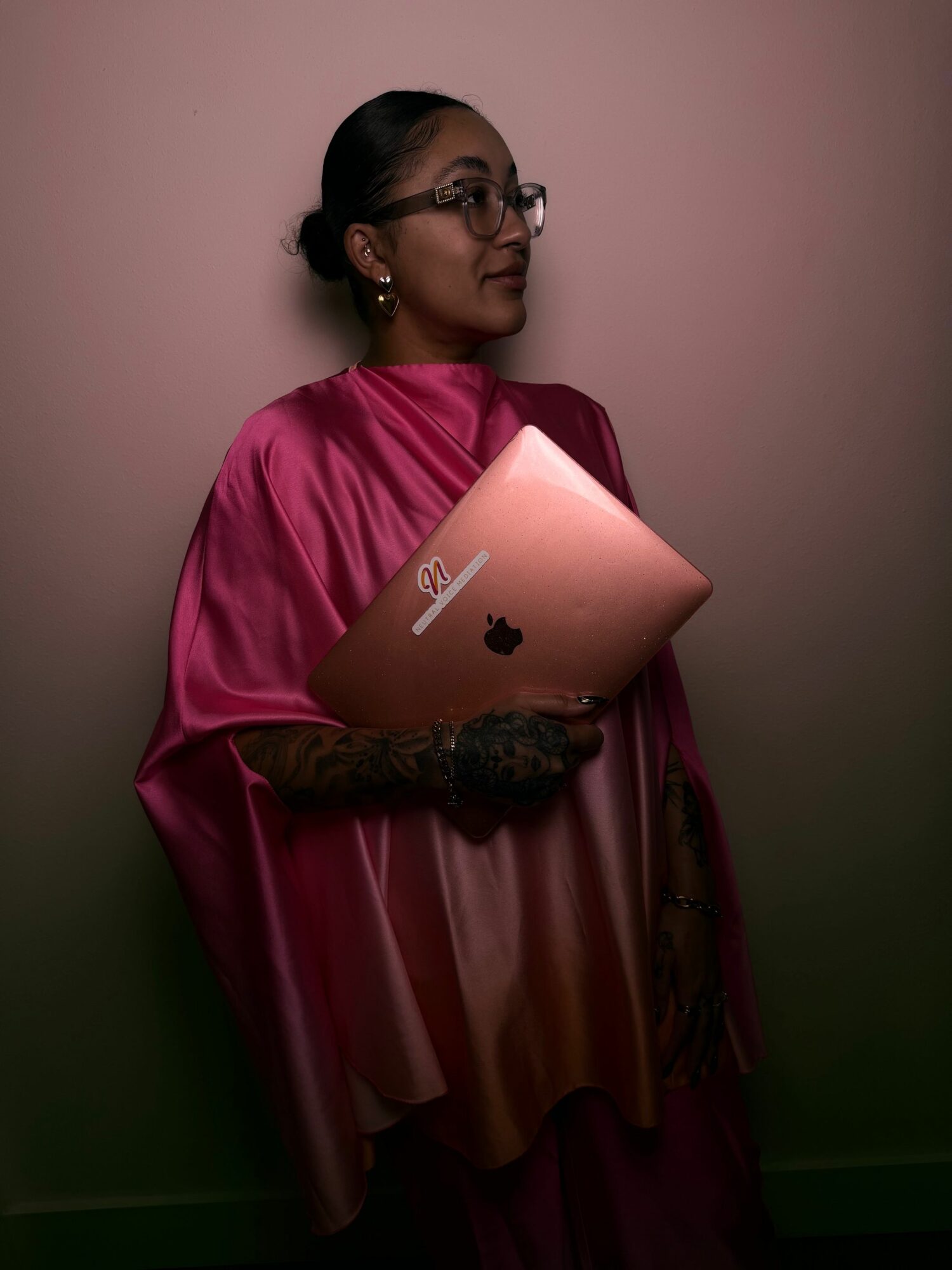 Woman in pink dress holding a pink laptop, standing against a plain wall, looking to the side.